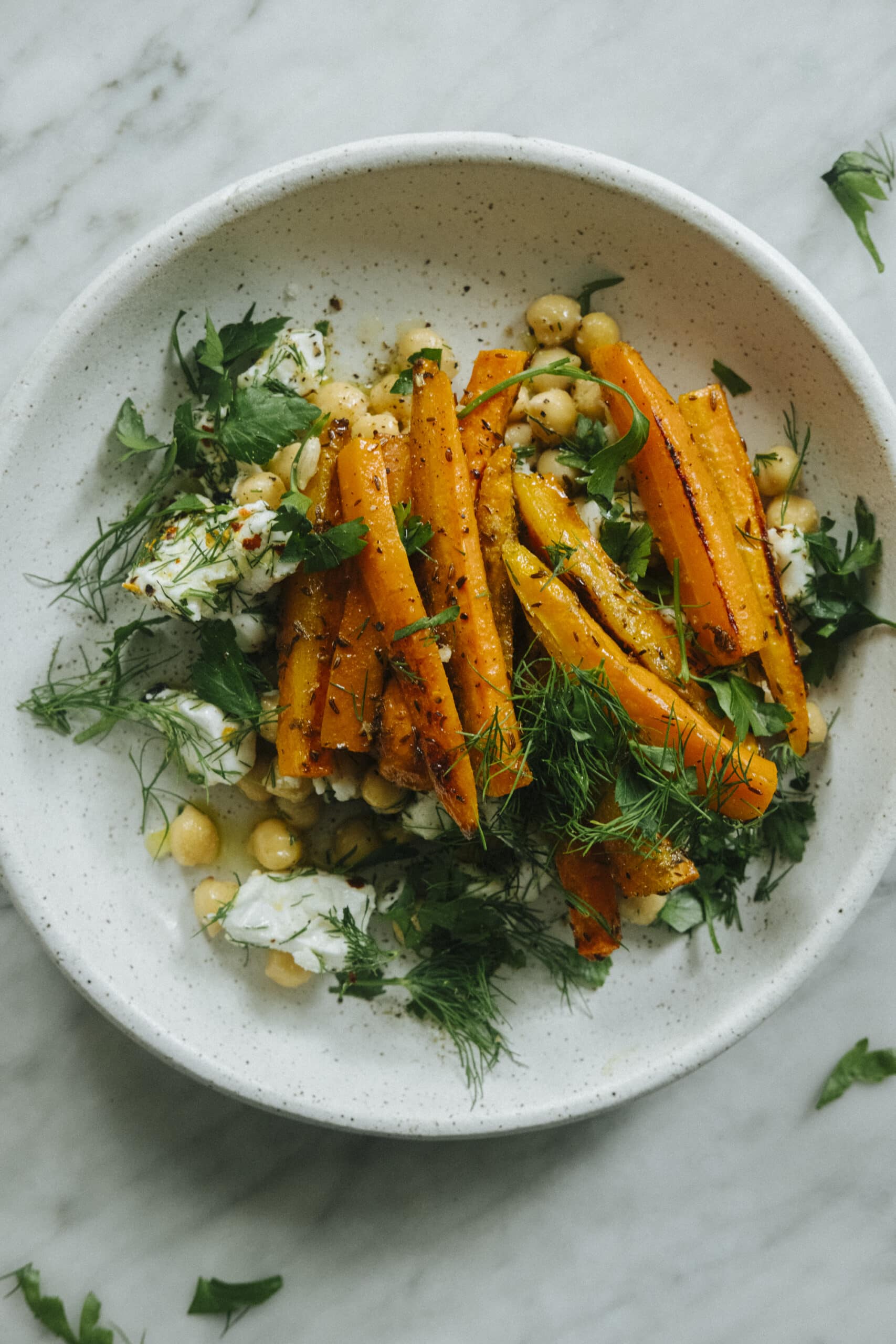 A white bowl filled with roasted carrots, chickpeas, fresh herbs, and a dollop of soft cheese on a marble surface.