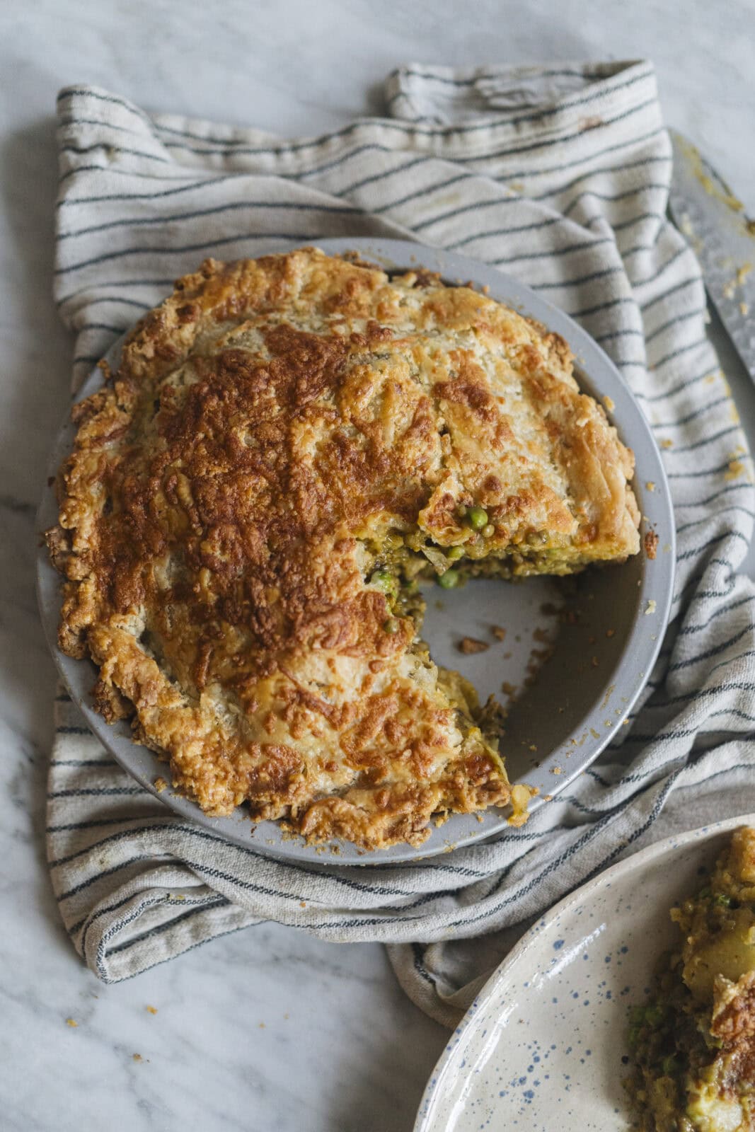 A round savory pie with a golden, flaky crust sits on a gray plate, missing one slice; it rests on a striped cloth over a marble surface. - Hetty Lui McKinnon