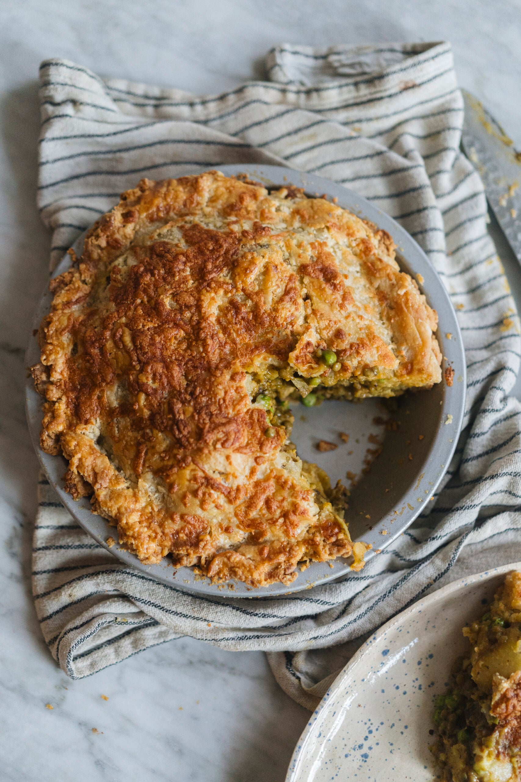 A round savory pie with a golden, flaky crust sits on a gray plate, missing one slice; it rests on a striped cloth over a marble surface. - Hetty Lui McKinnon