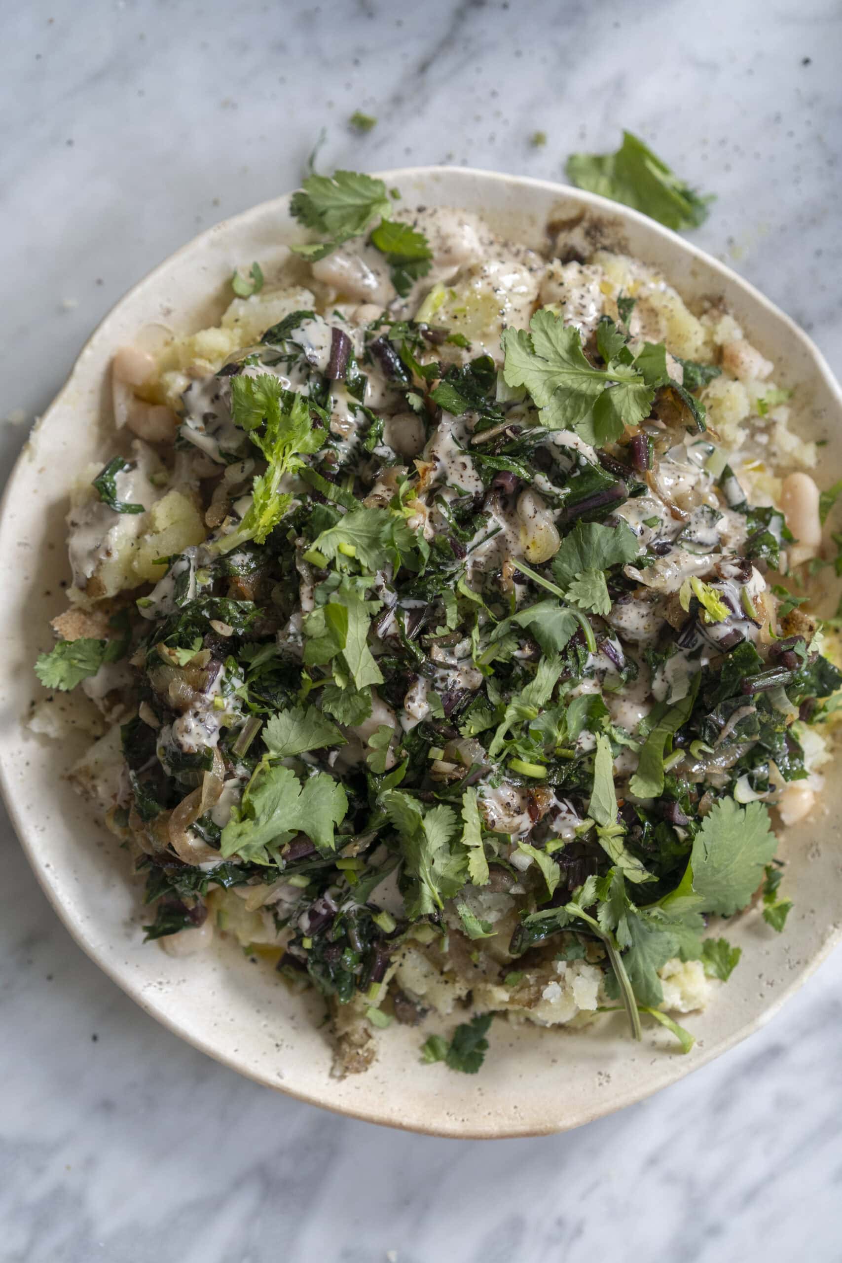 A plate of mashed potatoes topped with cooked greens, white beans, creamy sauce, and fresh chopped herbs on a marble surface.