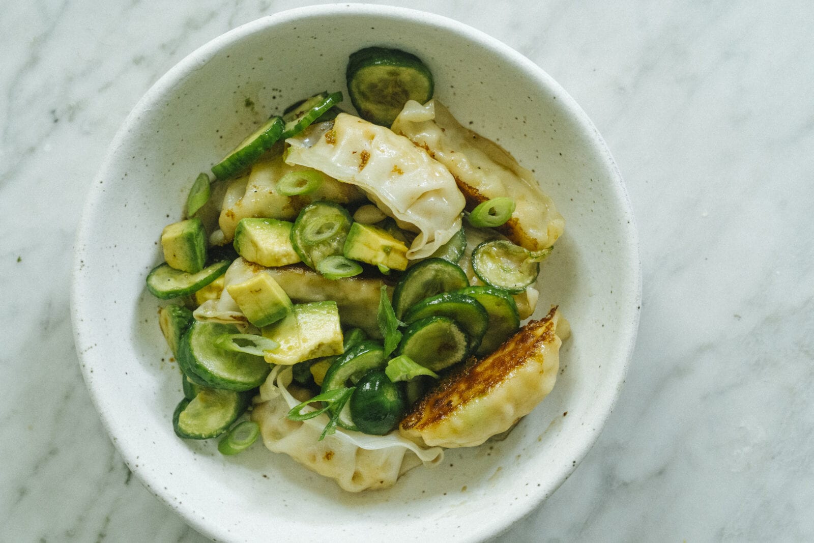 A white bowl filled with pan-fried dumplings, sliced cucumber, avocado, and green onions on a marble surface. - Hetty Lui McKinnon