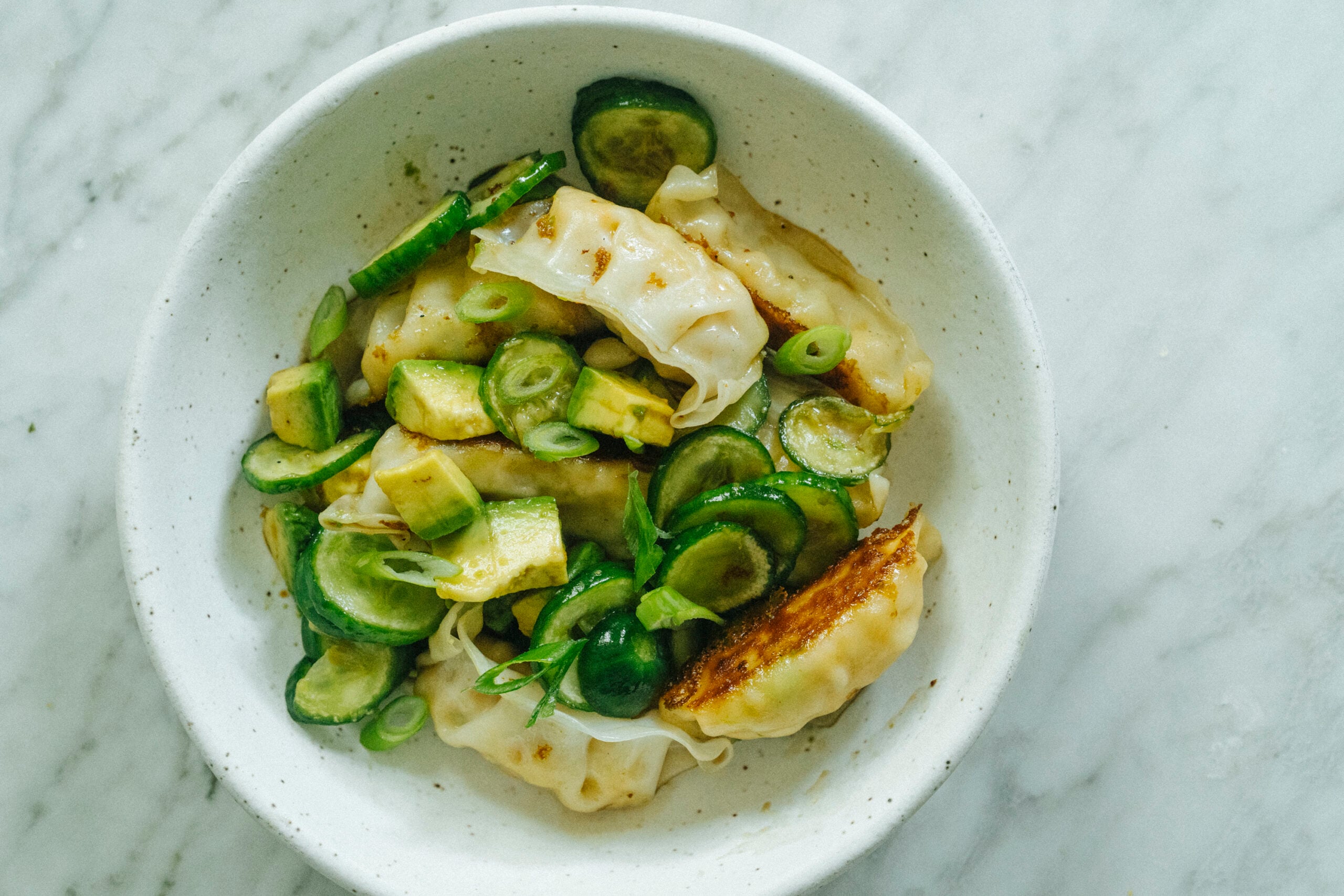 A white bowl filled with pan-fried dumplings, sliced cucumber, avocado, and green onions on a marble surface. - Hetty Lui McKinnon