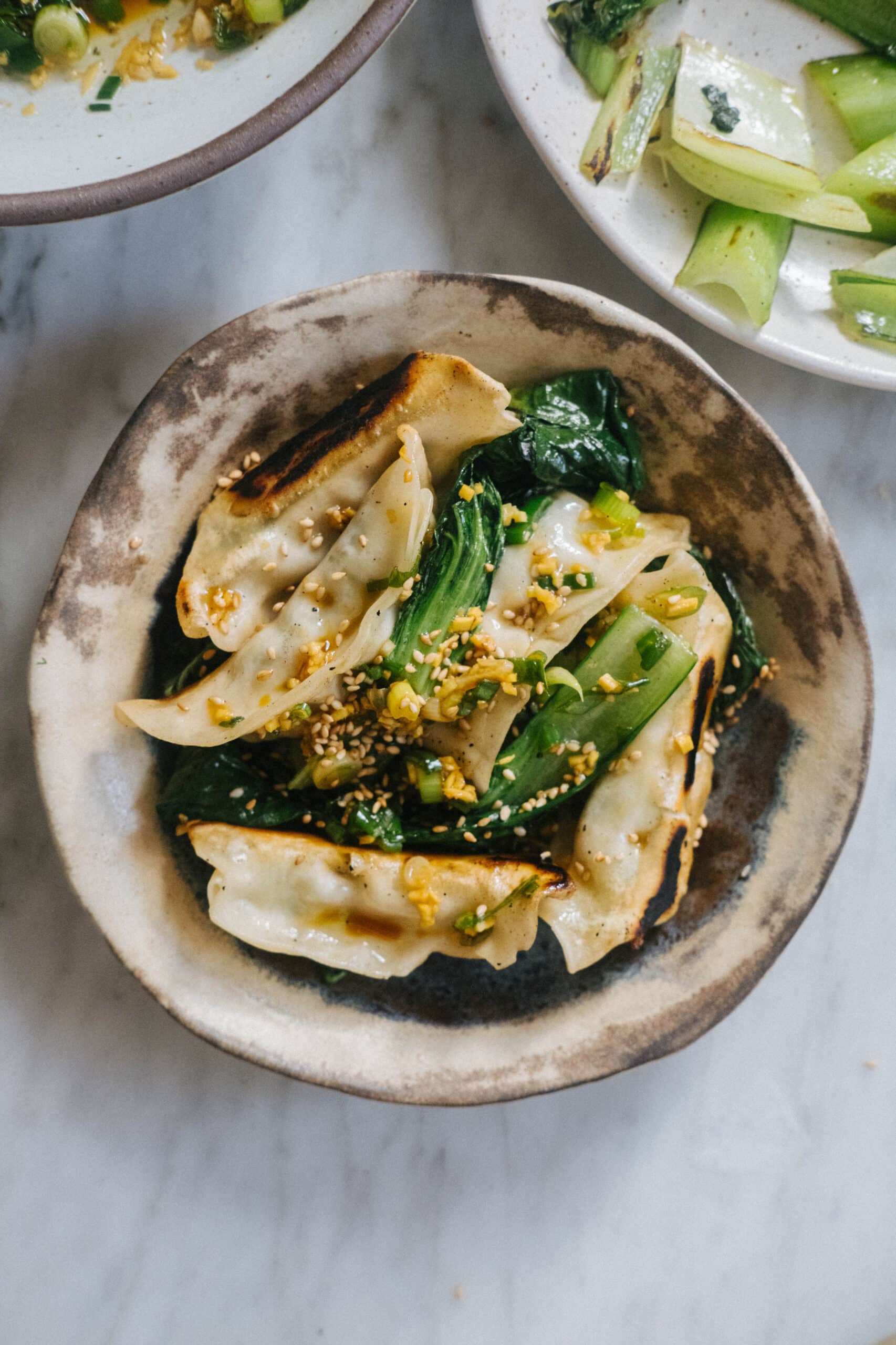 A ceramic bowl with pan-fried dumplings, sautéed greens, and a sprinkle of sesame seeds, placed on a marble surface.
