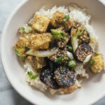 A bowl of white rice topped with sautéed eggplant, tofu cubes, sliced green onions, and sesame seeds.