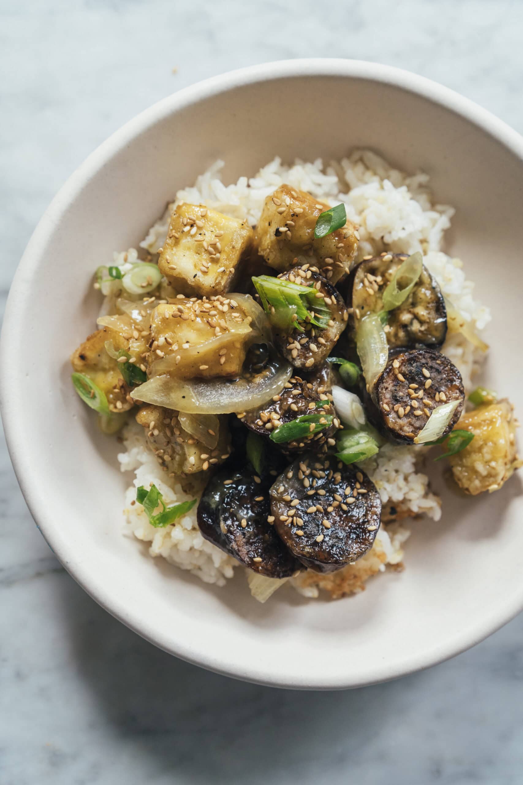A bowl of white rice topped with sautéed eggplant, tofu cubes, sliced green onions, and sesame seeds.