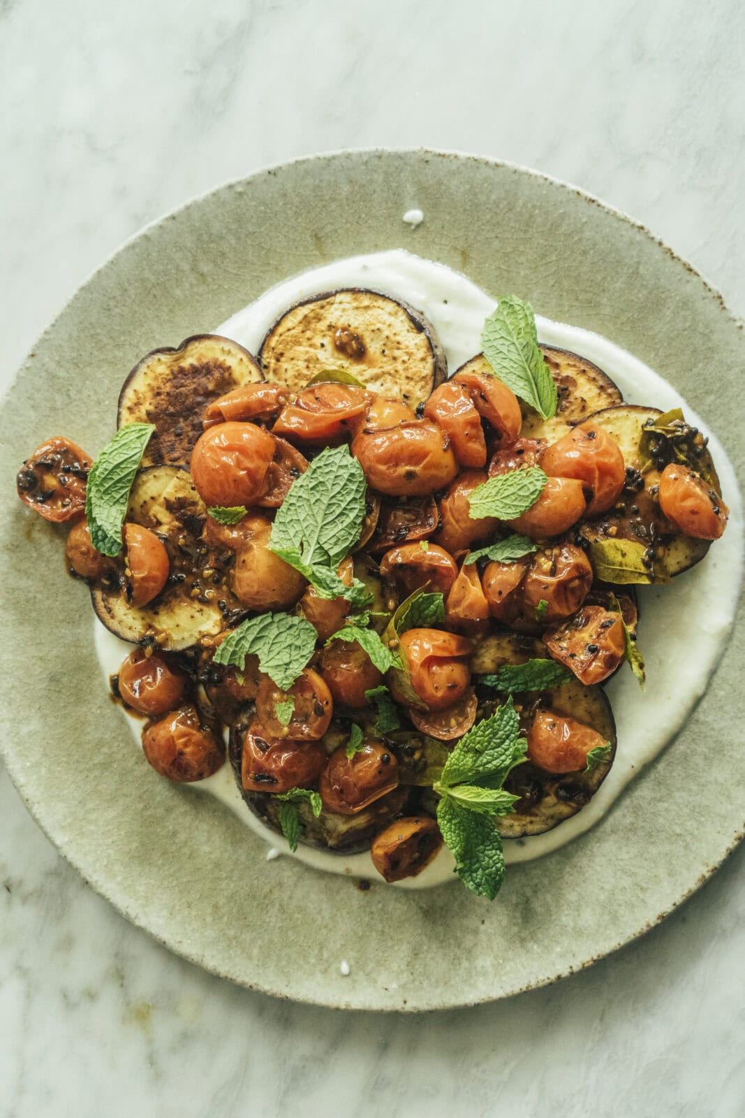 A plate of roasted eggplant and cherry tomatoes on a layer of yogurt, garnished with fresh mint leaves, on a marble surface. - Hetty Lui McKinnon