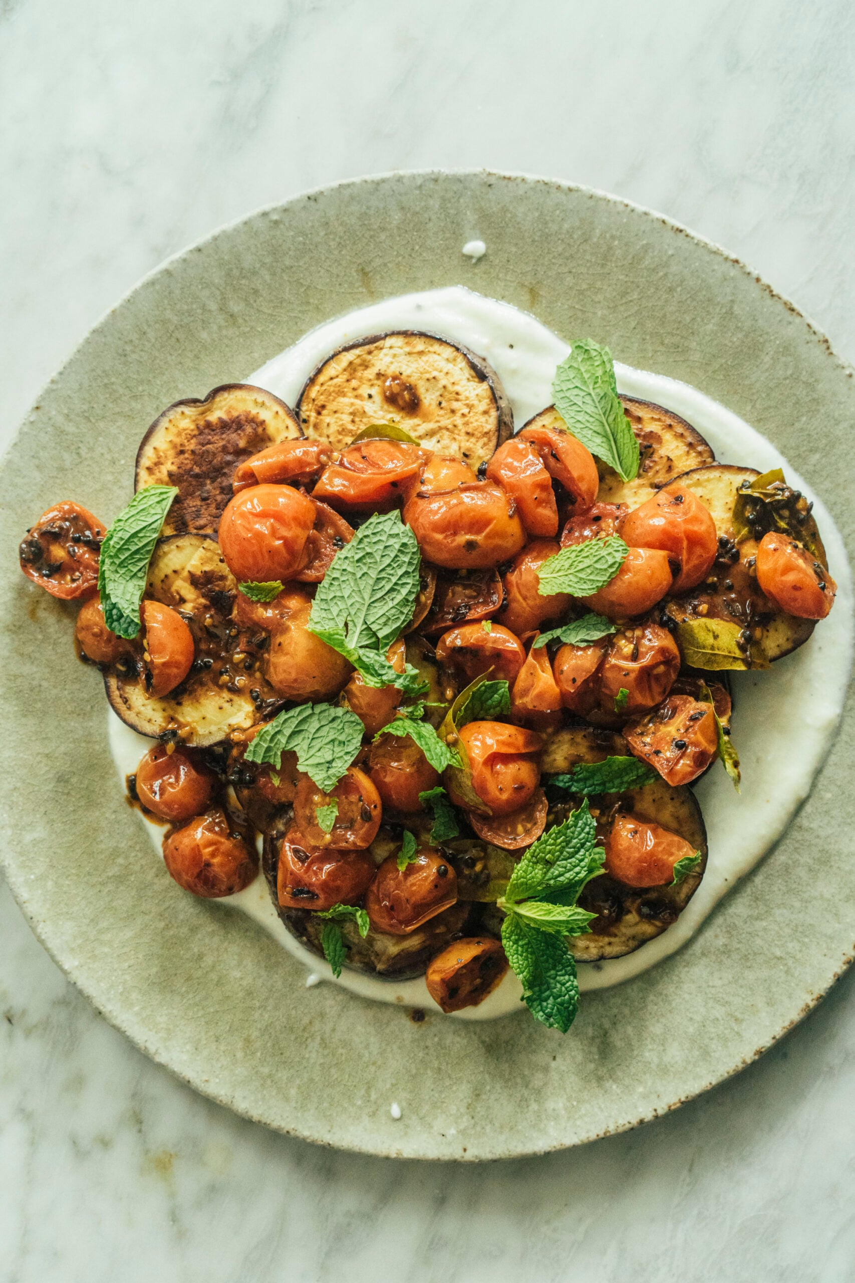 A plate of roasted eggplant and cherry tomatoes on a layer of yogurt, garnished with fresh mint leaves, on a marble surface. - Hetty Lui McKinnon