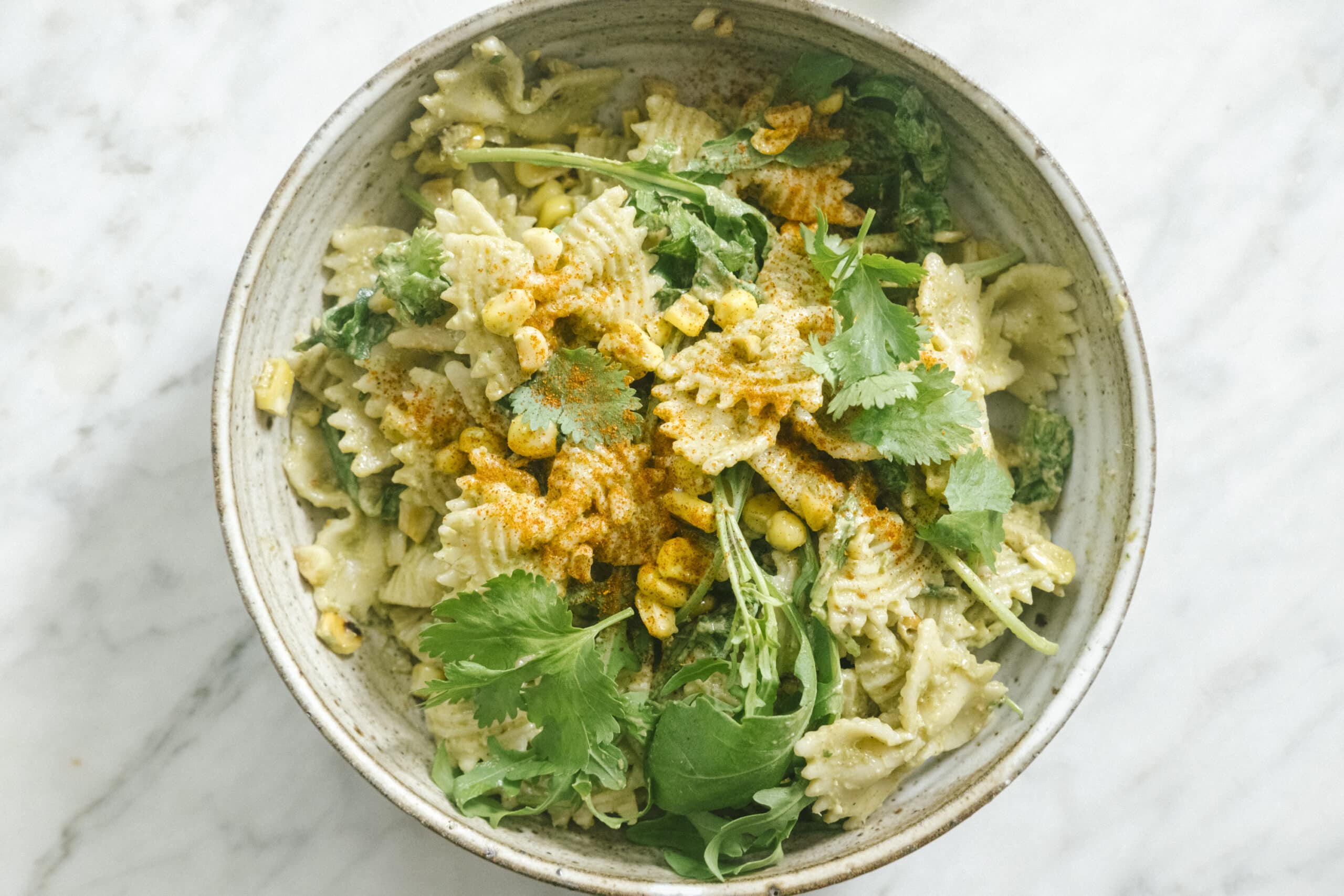 A bowl of farfalle pasta mixed with corn, leafy greens, cilantro, and a sprinkling of spices, served on a marble surface.