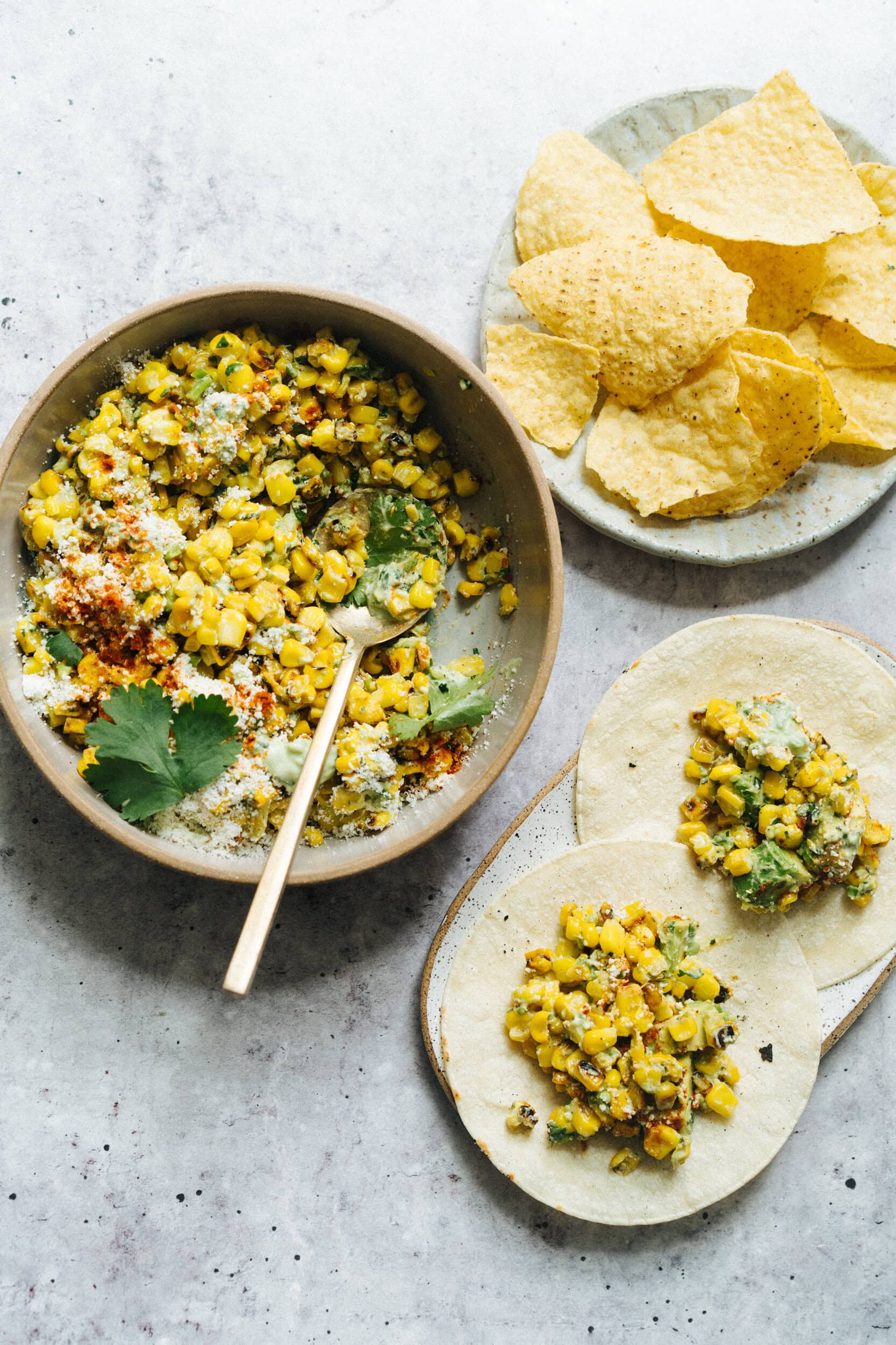 A bowl of seasoned grilled corn salad with a spoon, two corn tortillas topped with the salad, and a plate of tortilla chips on a light surface.