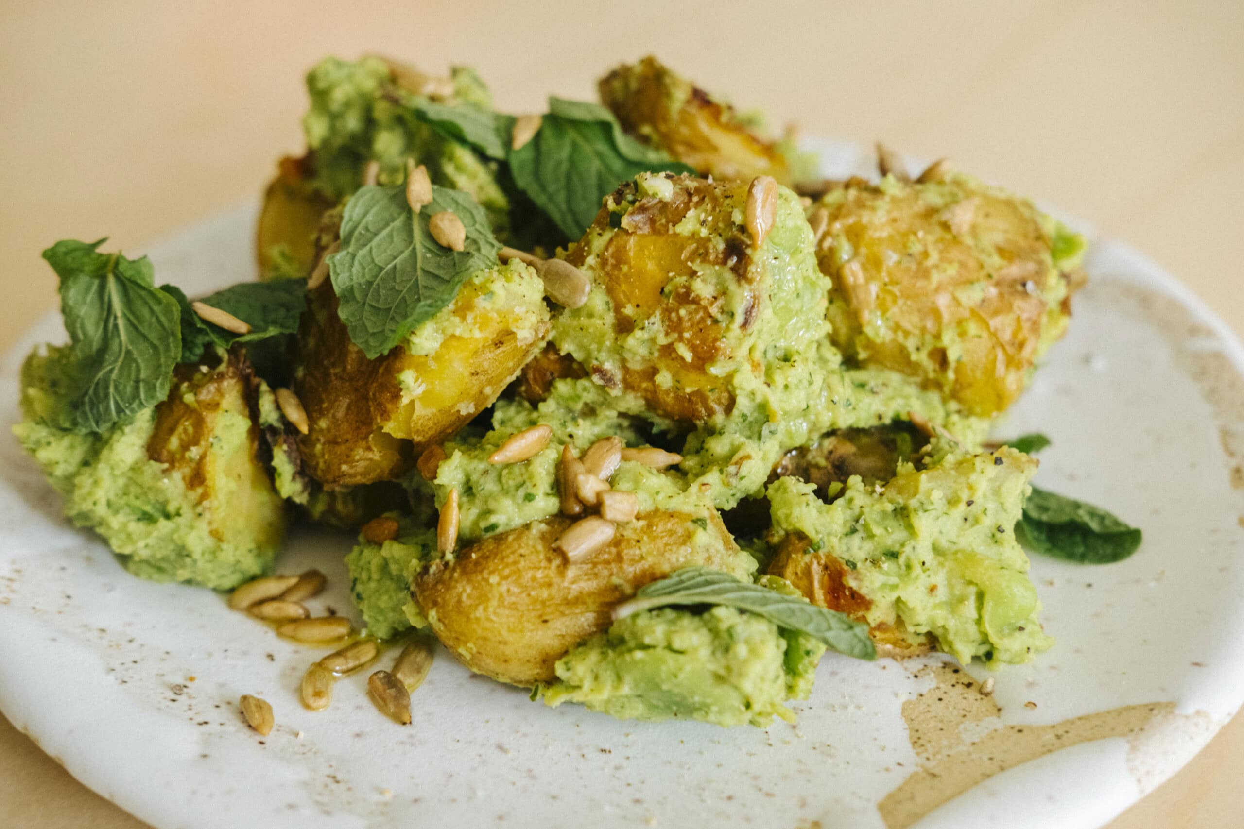 Roasted potato pieces with green herb sauce, garnished with fresh mint leaves and sunflower seeds on a white ceramic plate.