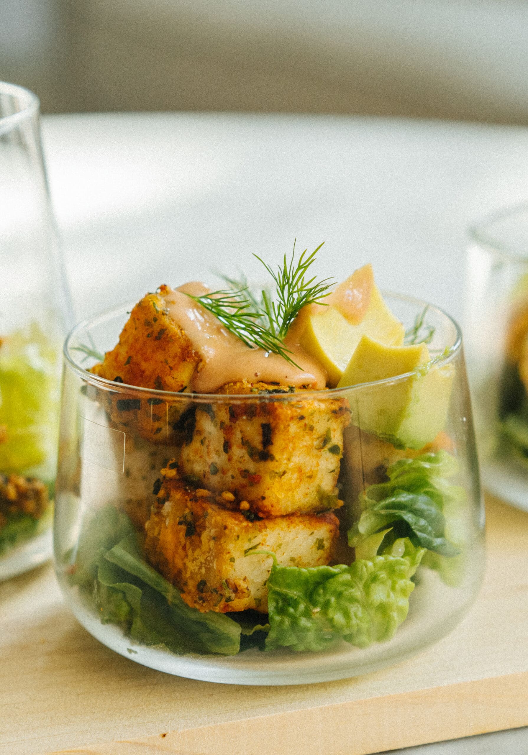 A clear glass cup filled with leafy greens, seasoned tofu cubes, avocado slices, fresh dill, and a dollop of creamy dressing.
