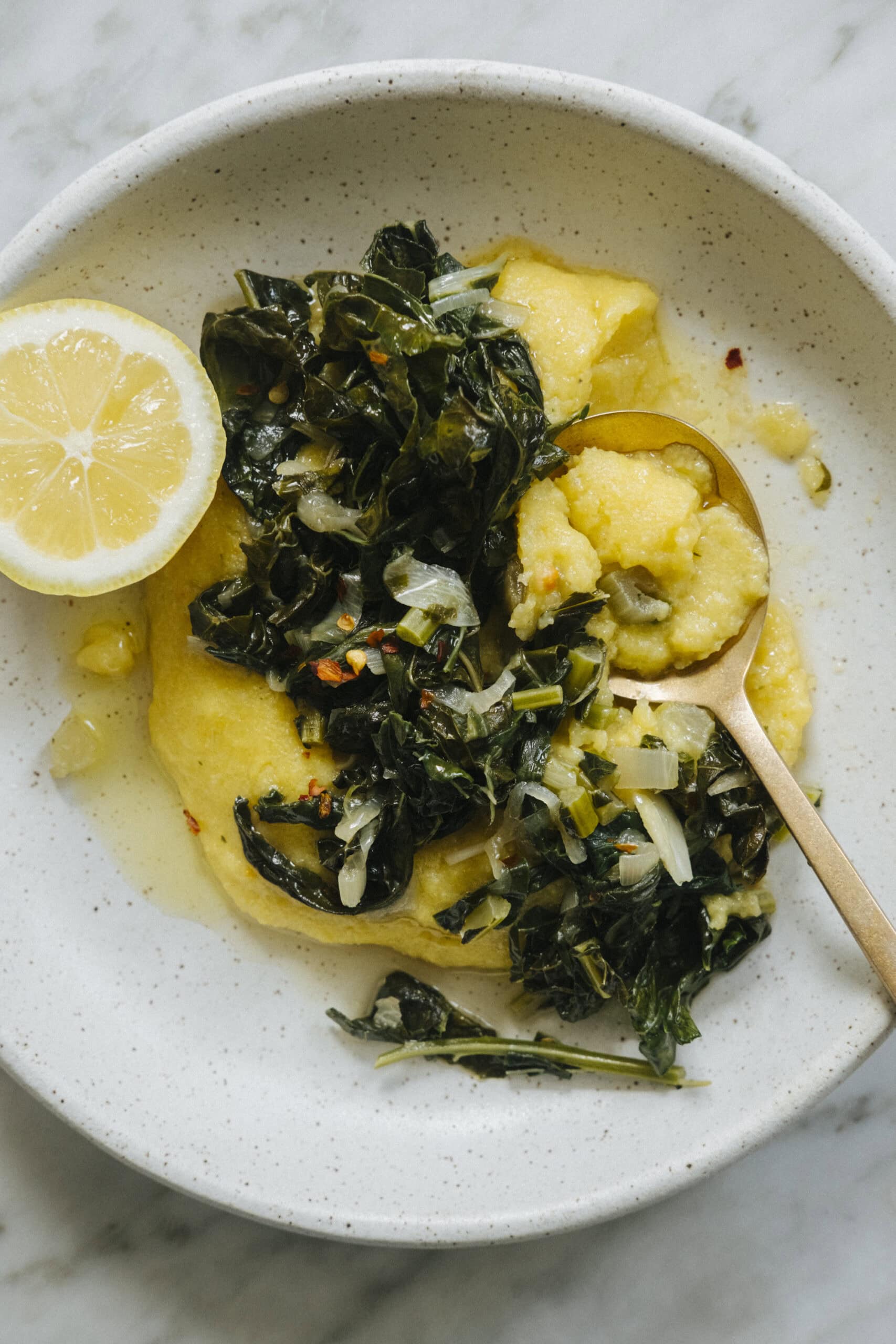 A plate of creamy polenta topped with sautéed greens and chopped onions, served with a lemon slice and a spoon.