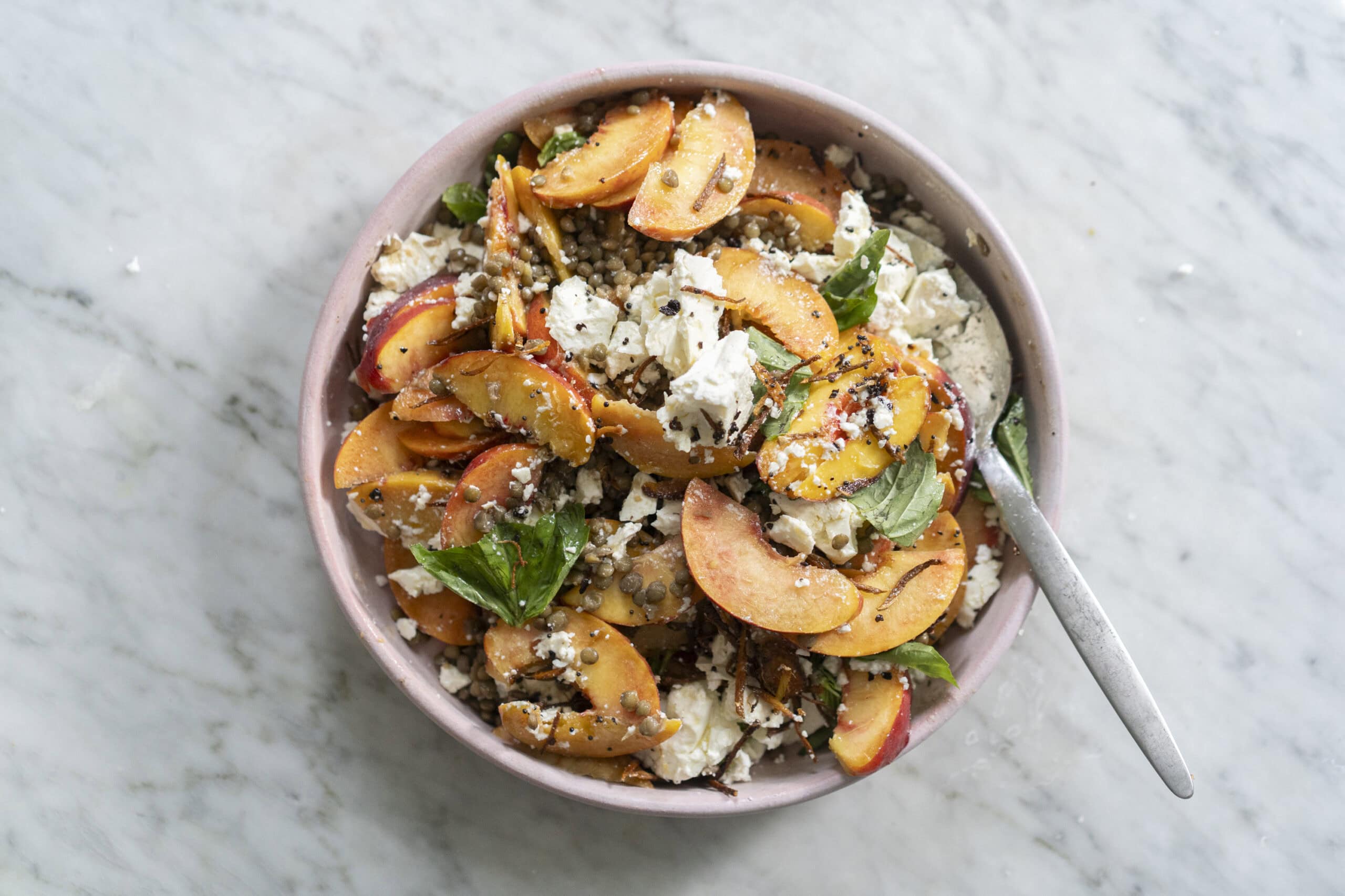 A bowl of salad with sliced peaches, crumbled cheese, greens, seeds, and grains on a marble surface with a spoon.