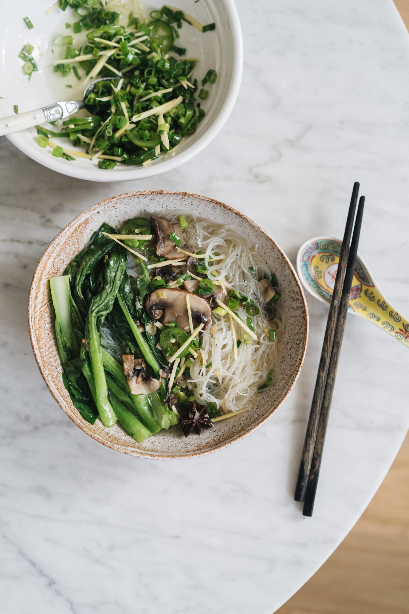 A bowl of rice noodle soup with bok choy, mushrooms, and herbs sits on a marble table beside a bowl of chopped green onions and a pair of chopsticks with a ceramic spoon.