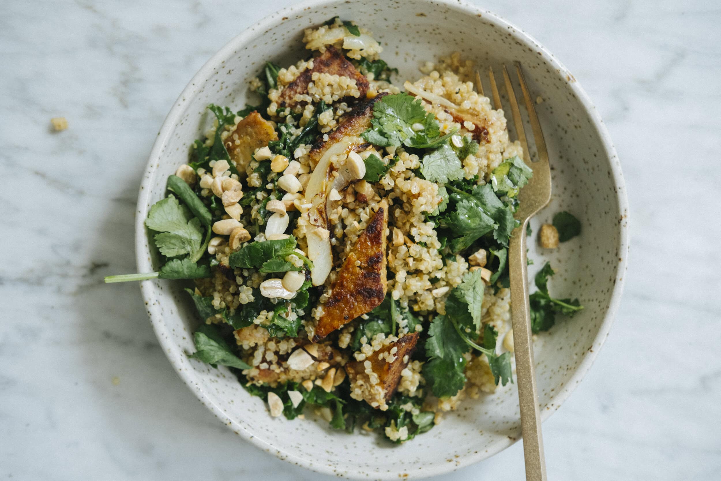 A bowl of quinoa salad with greens, chopped nuts, grilled chicken pieces, and a fork on a marble surface.