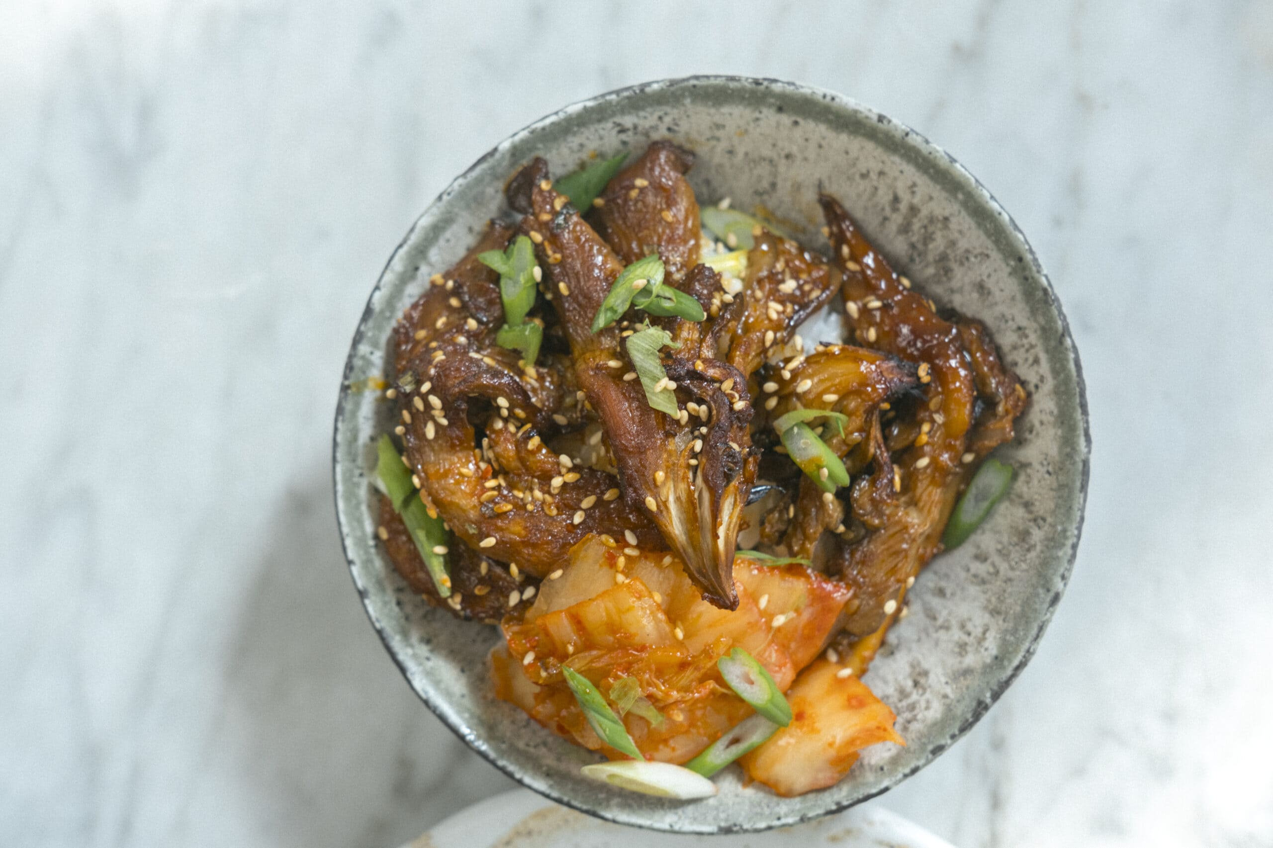A bowl of rice topped with glazed shredded meat, kimchi, sliced green onions, and sesame seeds on a light marble surface.