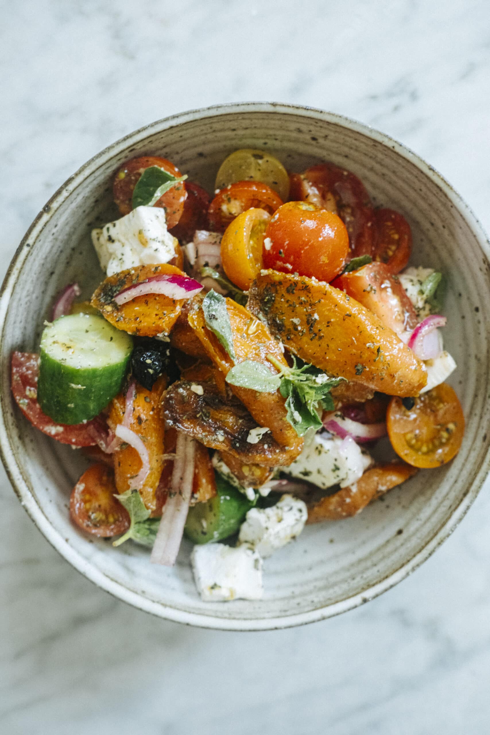 A bowl of salad containing cherry tomatoes, sliced cucumbers, red onions, roasted carrots, feta cheese, herbs, and a light dressing on a marble surface.