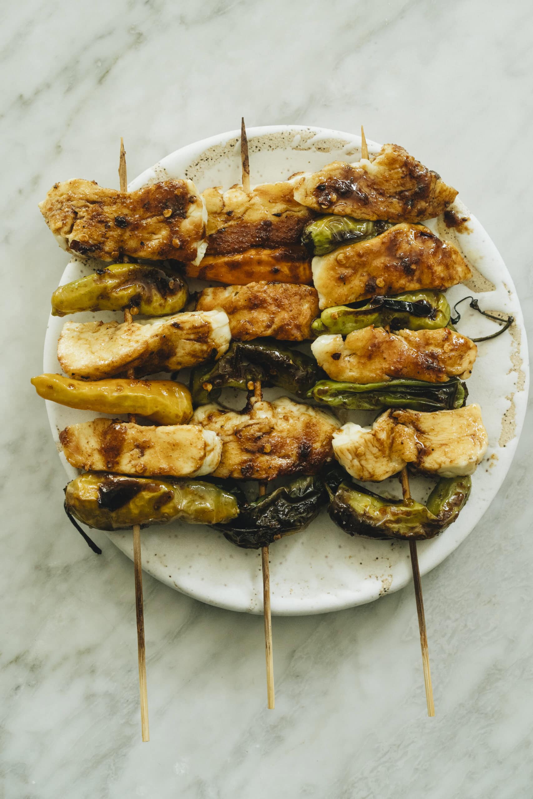 Three grilled skewers with pieces of chicken and green peppers arranged on a round white plate atop a marble surface. - Hetty Lui McKinnon
