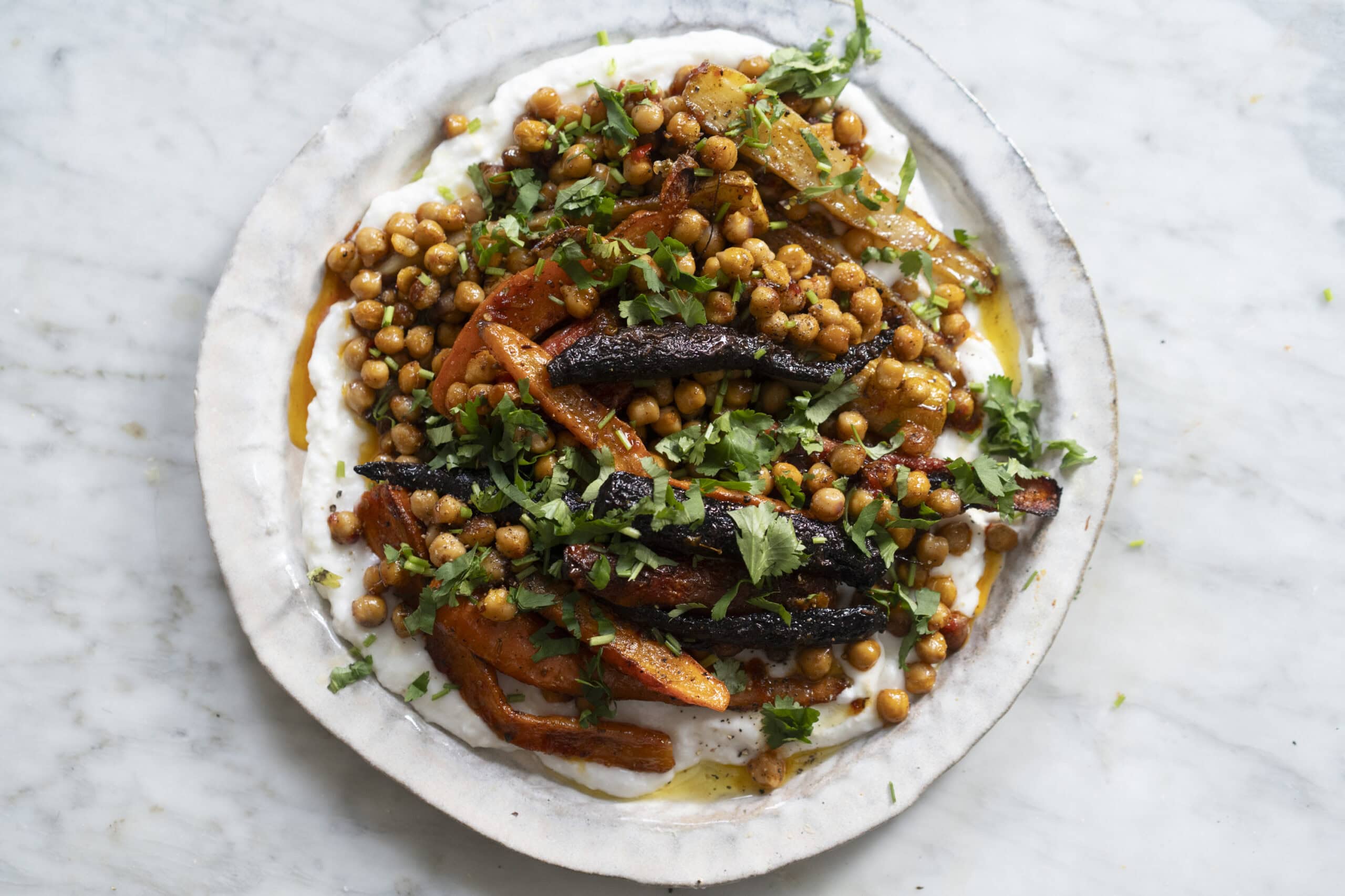 A plate of roasted carrots, chickpeas, and onions served over a creamy white sauce, garnished with chopped fresh herbs.