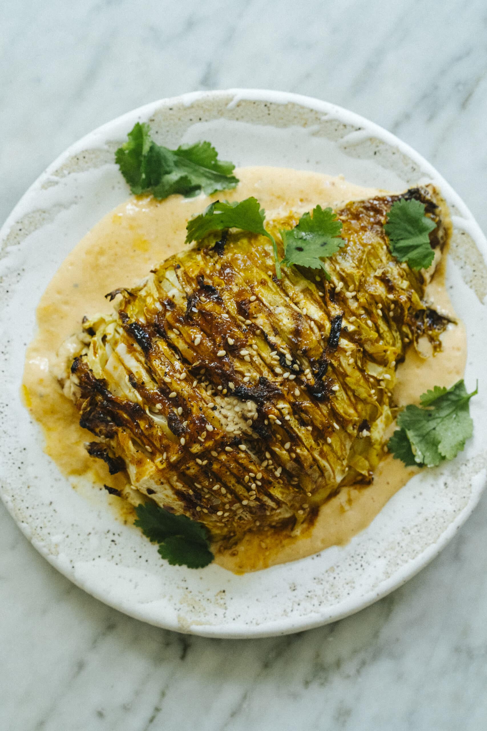 Roasted cabbage wedge drizzled with sauce, topped with sesame seeds and cilantro on a white plate. - Hetty Lui McKinnon