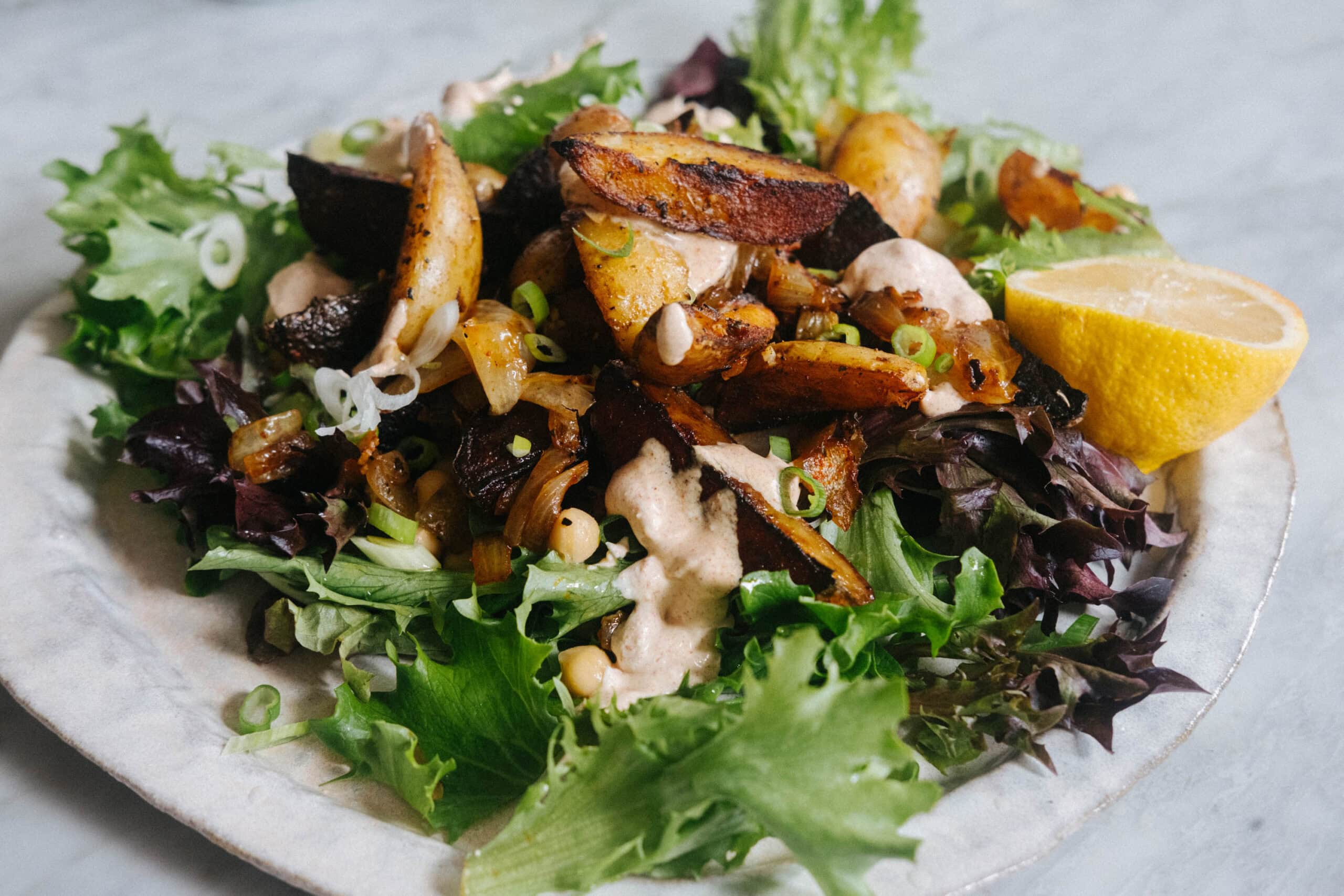 A plate of mixed salad greens topped with roasted potatoes, chickpeas, sliced green onions, a creamy dressing, and a lemon wedge on the side.