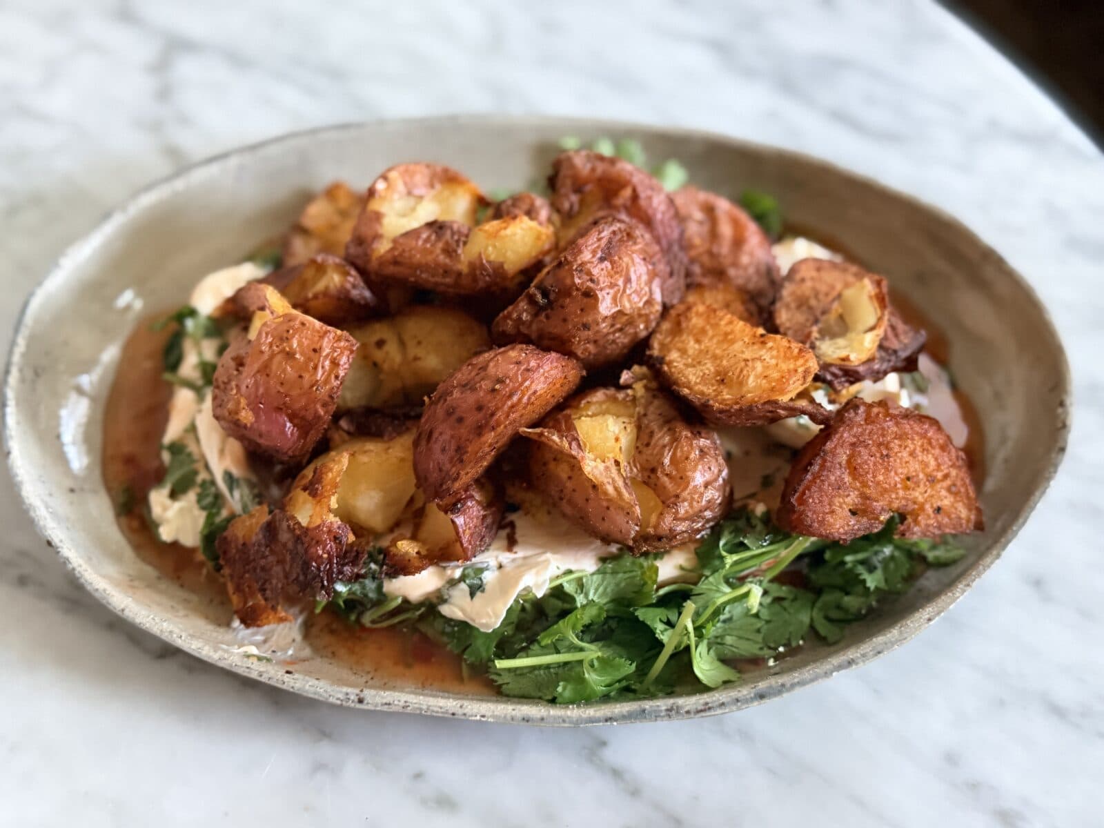 A ceramic plate with crispy roasted potatoes on a bed of greens and creamy sauce, served on a marble surface.