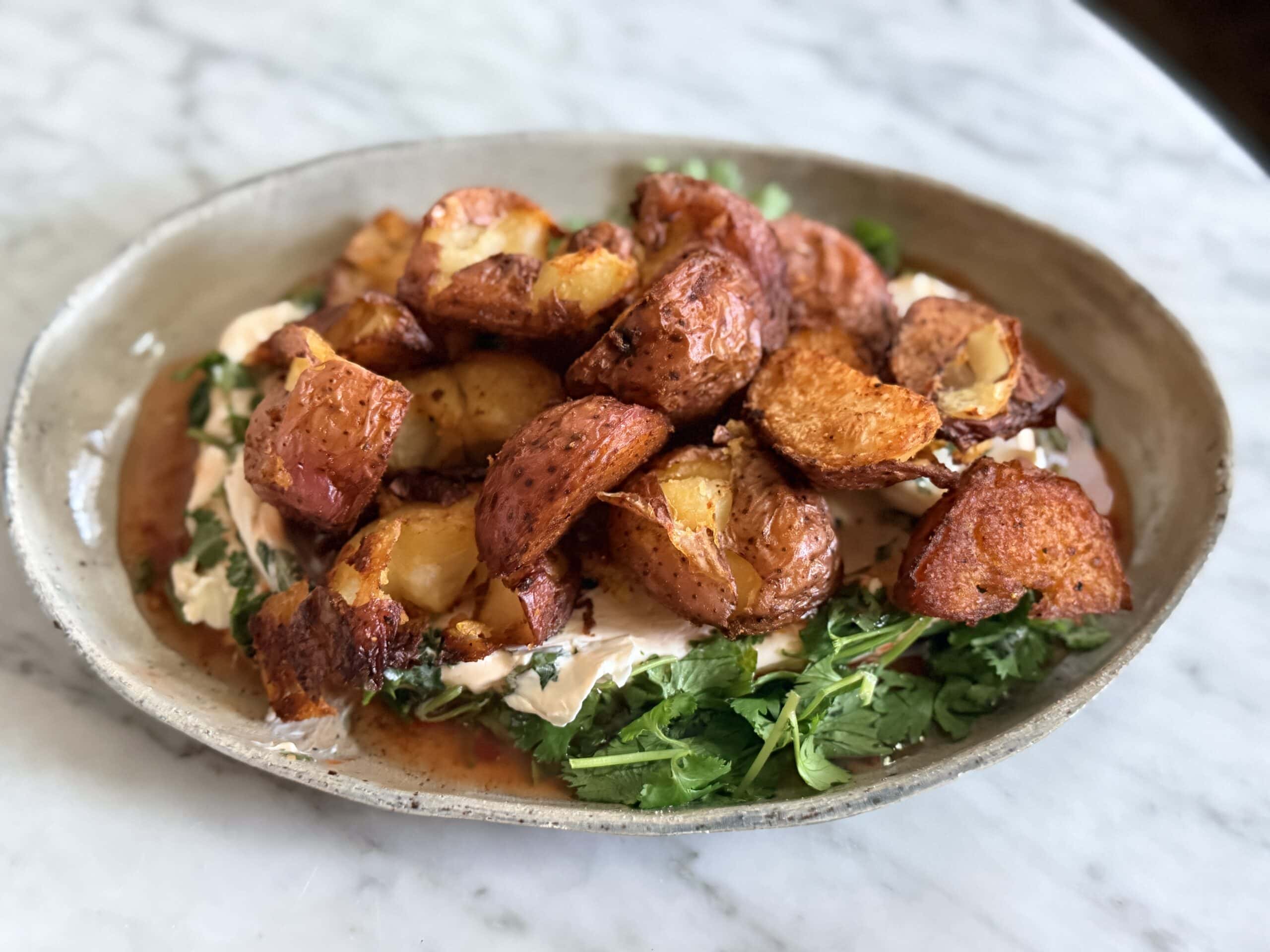 A ceramic plate with crispy roasted potatoes on a bed of greens and creamy sauce, served on a marble surface.