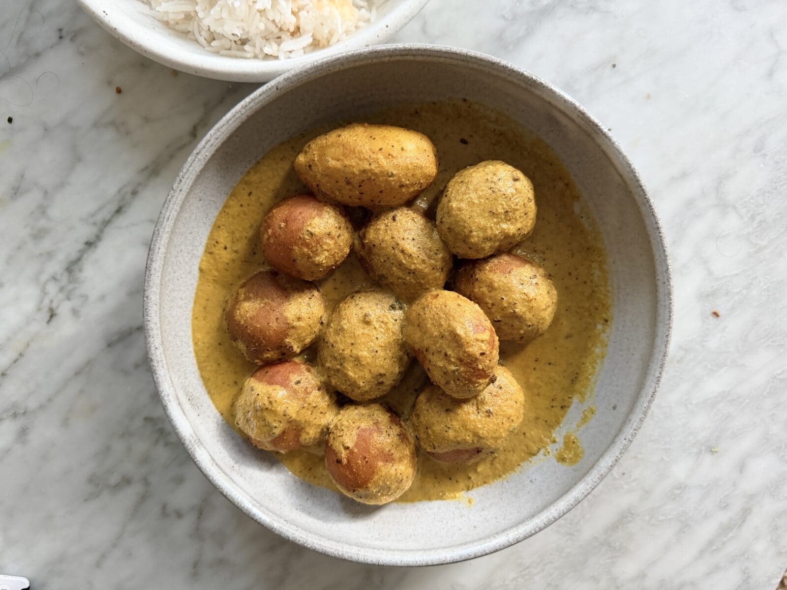 A bowl of small potatoes in a yellow curry sauce sits on a marble surface, with a partial bowl of white rice visible in the corner. - Hetty Lui McKinnon