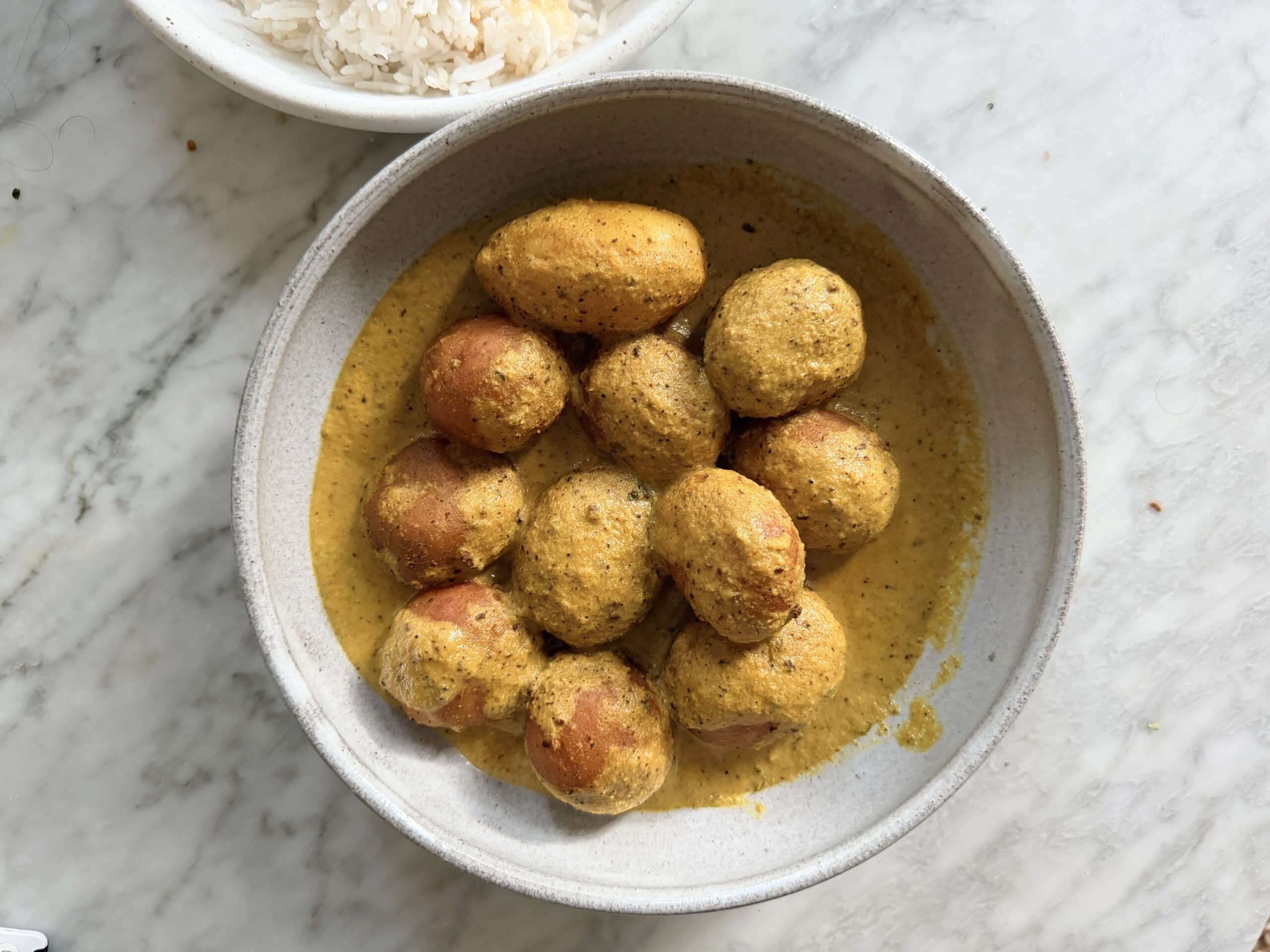A bowl of small potatoes in a yellow curry sauce sits on a marble surface, with a partial bowl of white rice visible in the corner. - Hetty Lui McKinnon