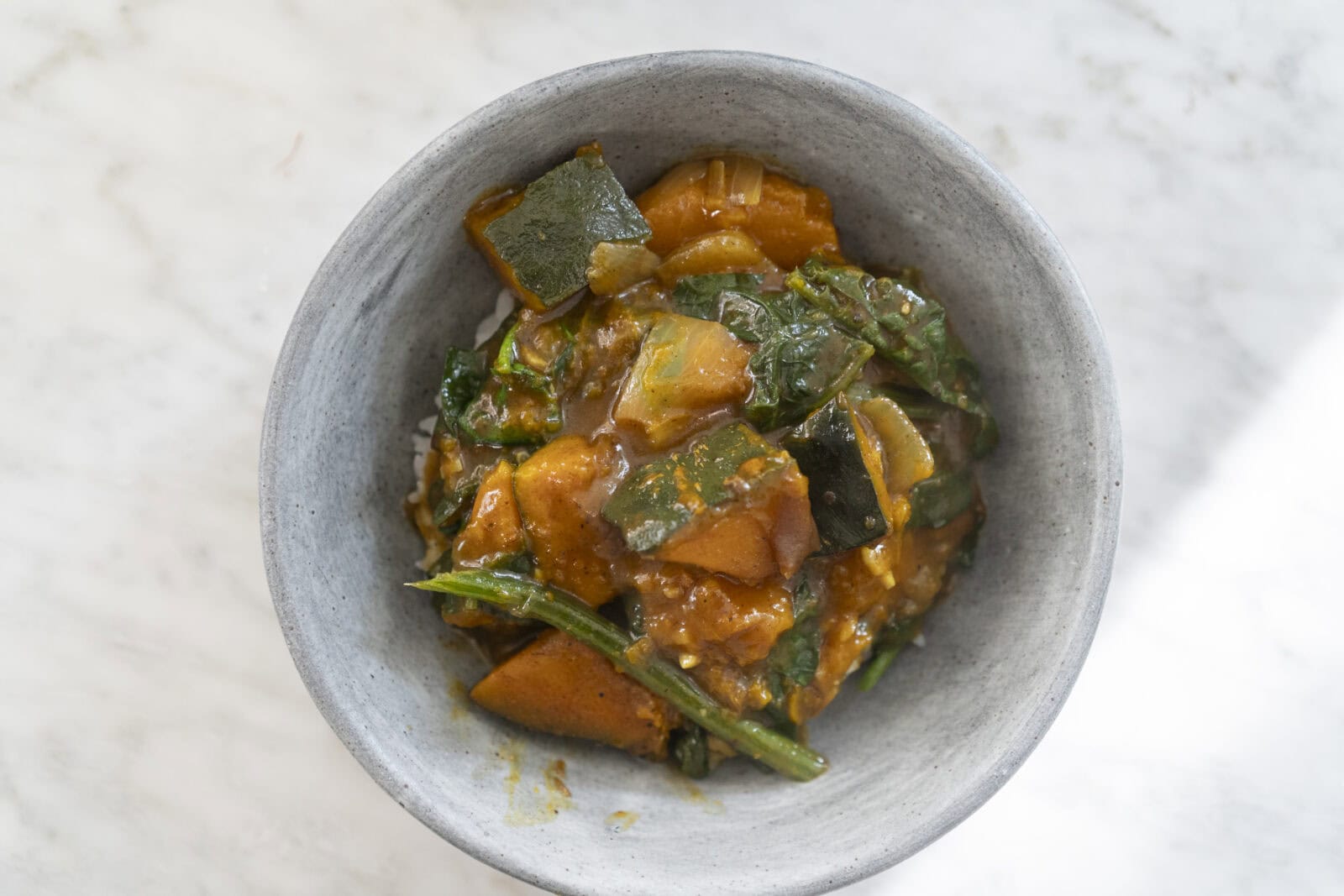 A bowl of vegetable curry featuring chunks of squash, leafy greens, and a thick orange sauce served over rice.