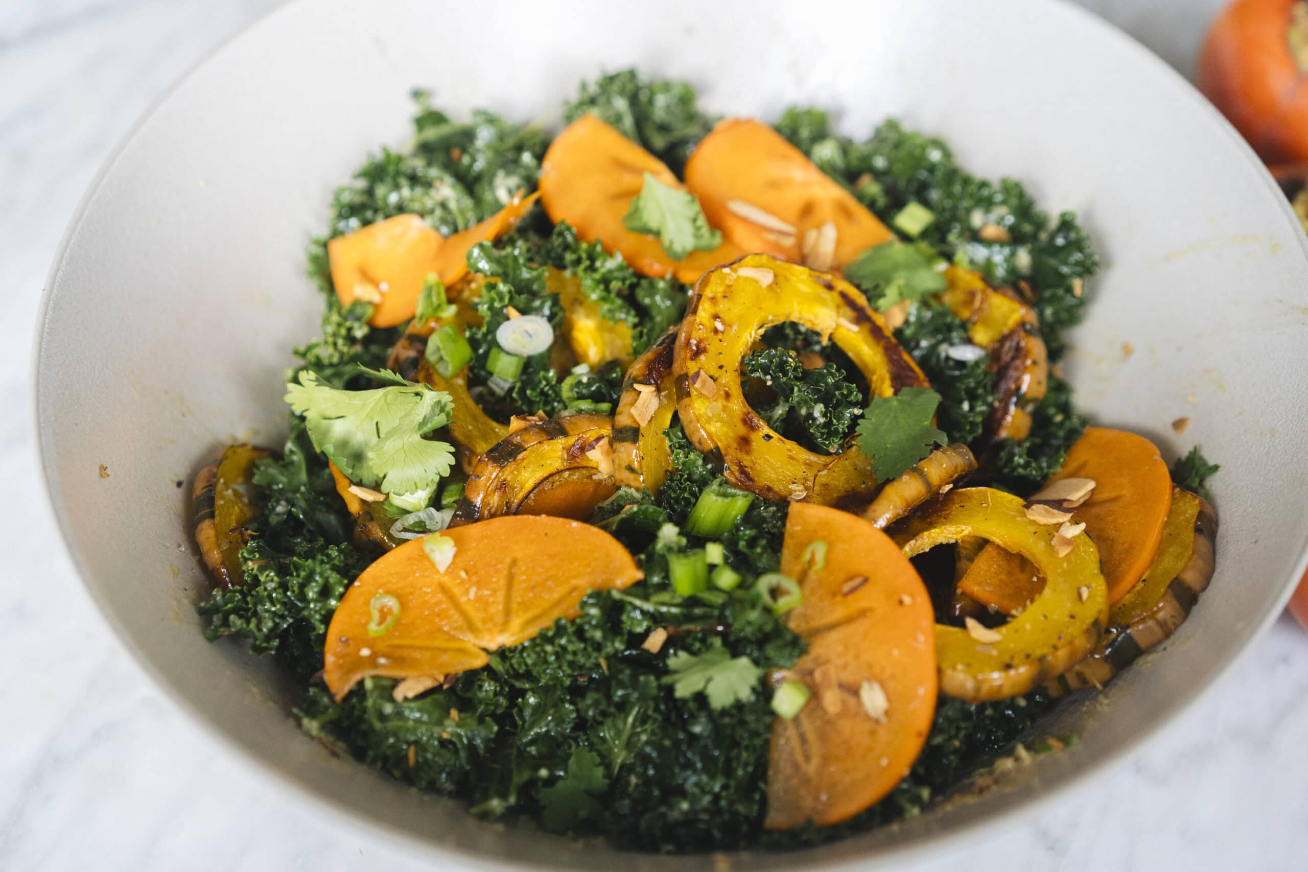 A salad with kale, sliced roasted squash, orange persimmons, green onions, cilantro, and sunflower seeds in a white bowl.