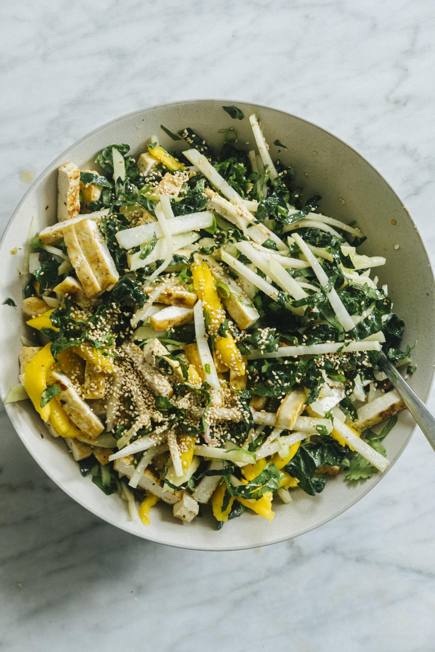 A bowl of salad with sliced tofu, yellow bell peppers, kale, jicama, and sesame seeds on a marble surface.