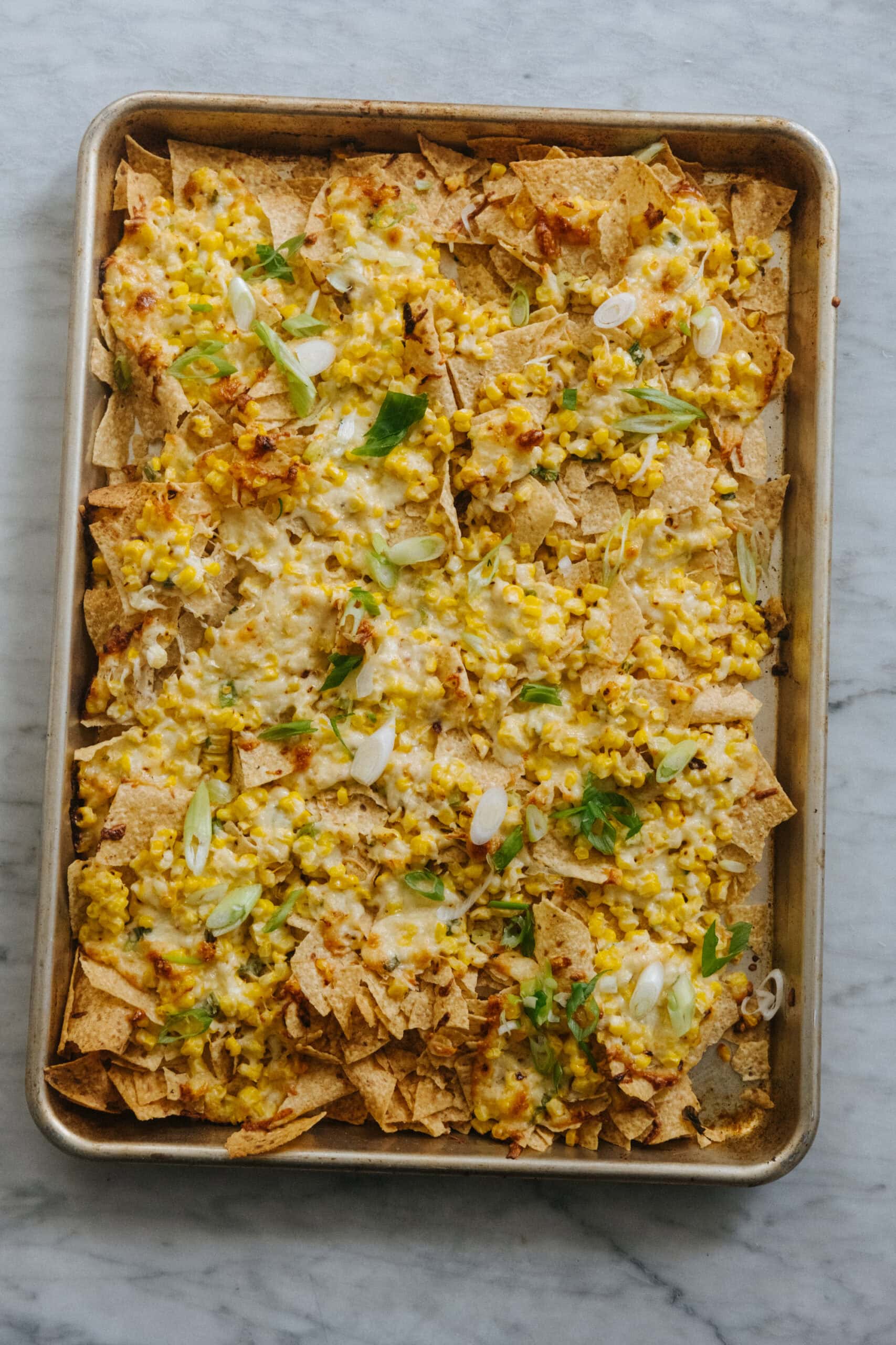 A sheet pan of nachos topped with melted cheese, corn, sliced green onions, and seasonings on a marble surface.