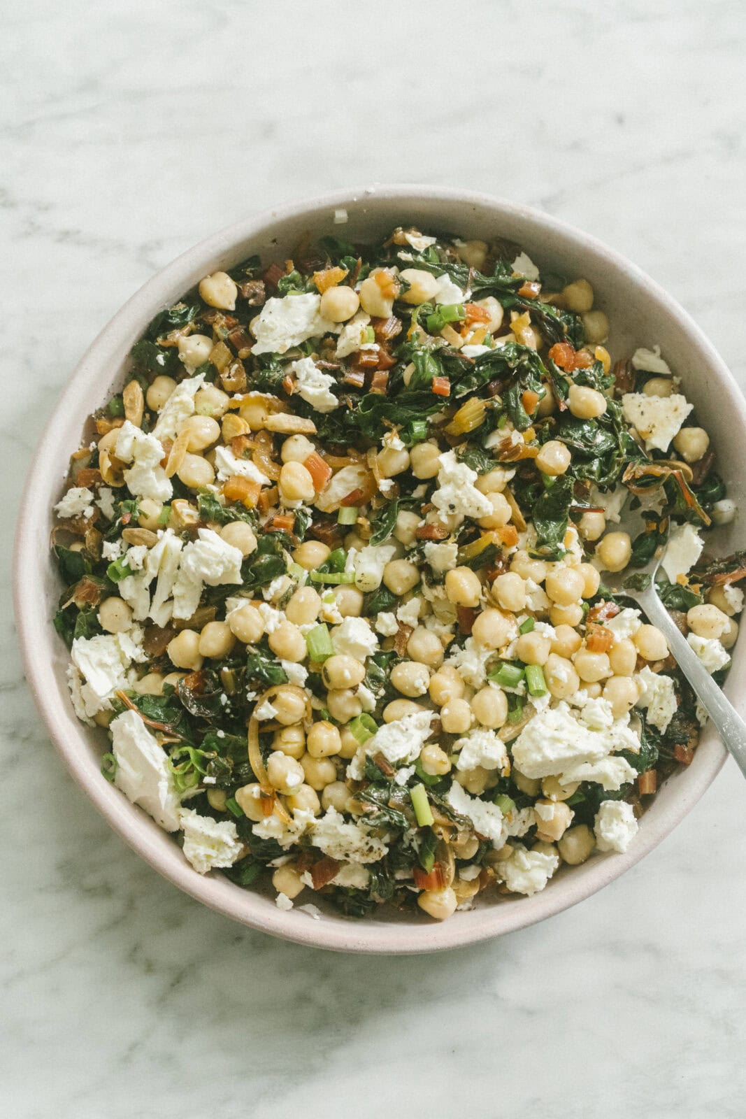 A bowl of chickpea salad with chopped greens, crumbled feta cheese, and diced vegetables on a marble surface.