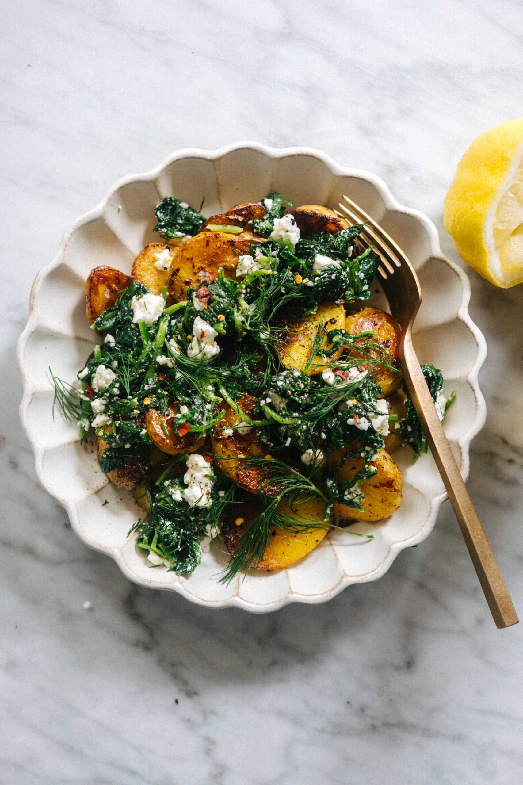 A ceramic bowl with roasted potato slices, wilted greens, fresh dill, and crumbled white cheese, placed on a marble surface with a lemon wedge nearby.