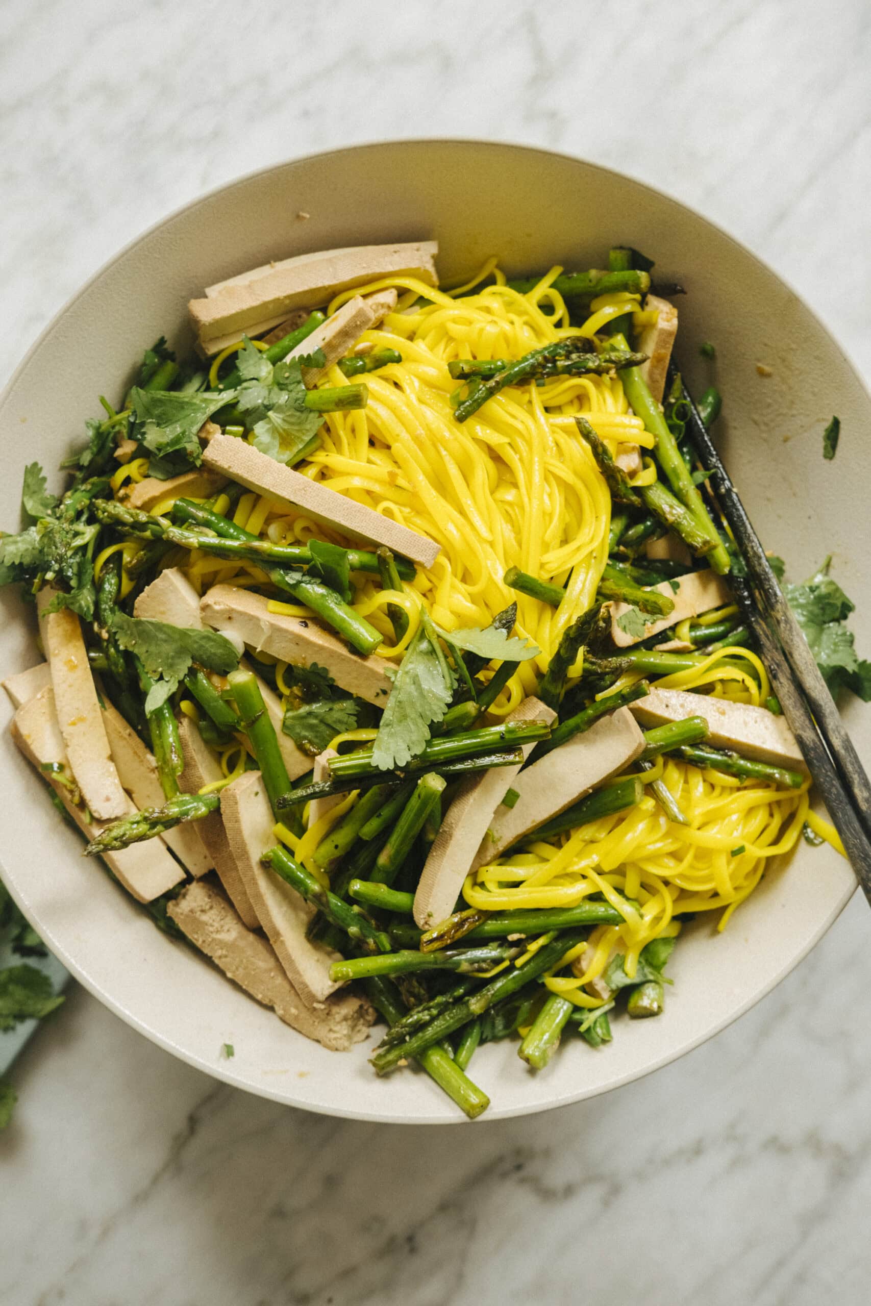 A bowl of yellow noodles mixed with sliced tofu, asparagus, and fresh cilantro, with chopsticks resting on the edge.