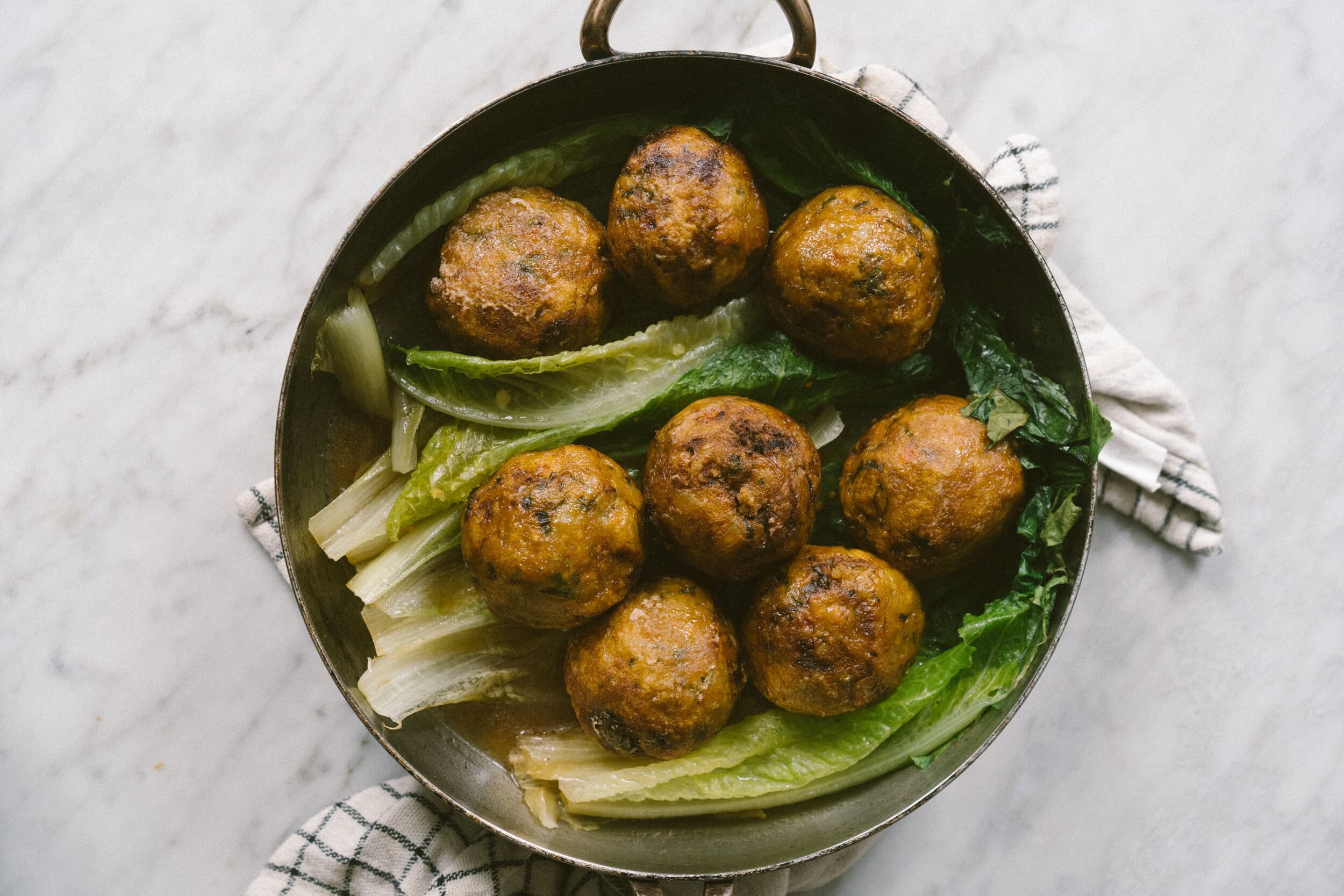A round metal pan containing eight browned meatballs placed on a bed of leafy greens, with a striped cloth partially visible underneath.