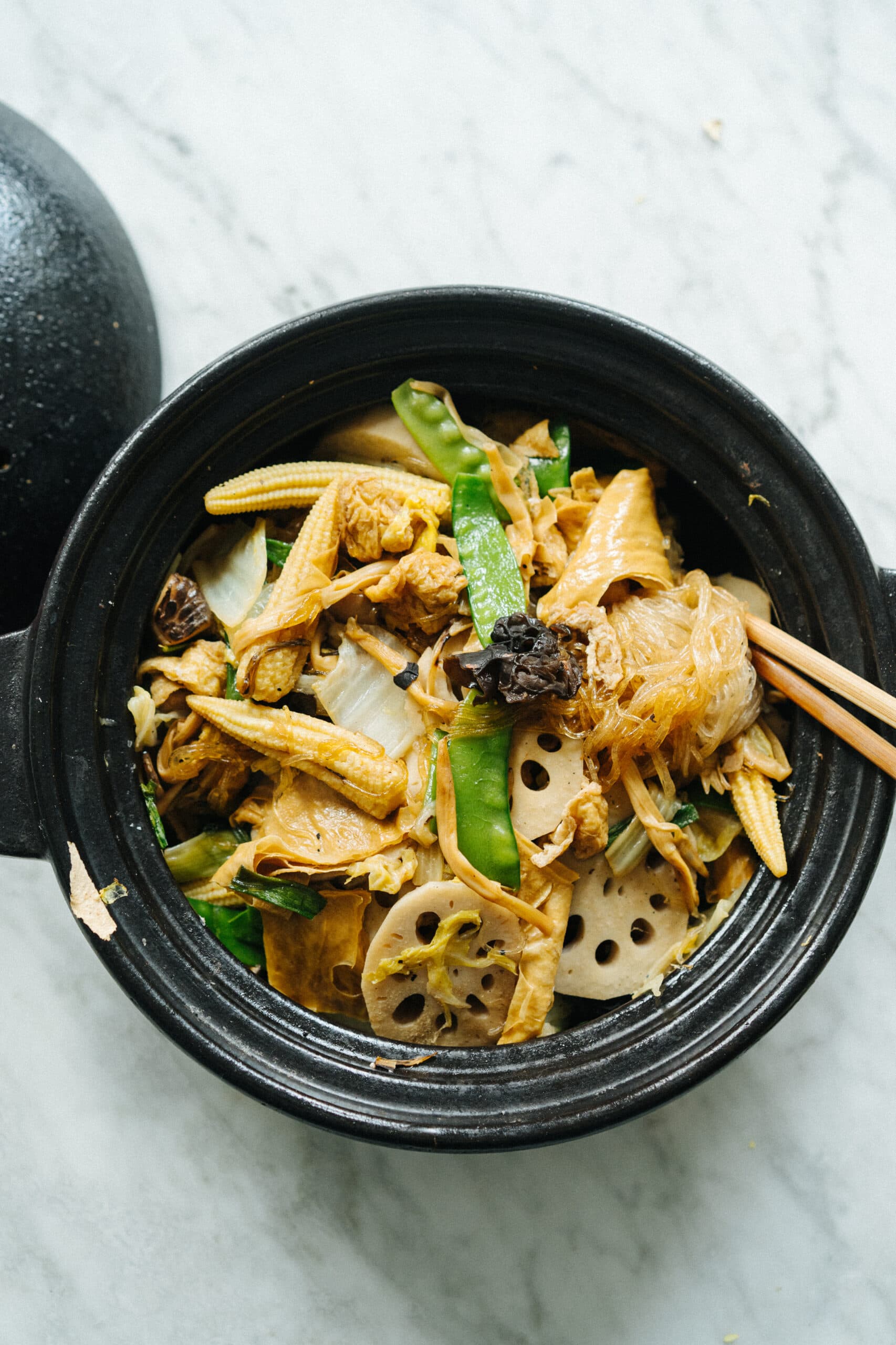 A black bowl contains a mixed vegetable stir-fry with lotus root, baby corn, snow peas, mushrooms, and noodles. Chopsticks rest on the side of the bowl.