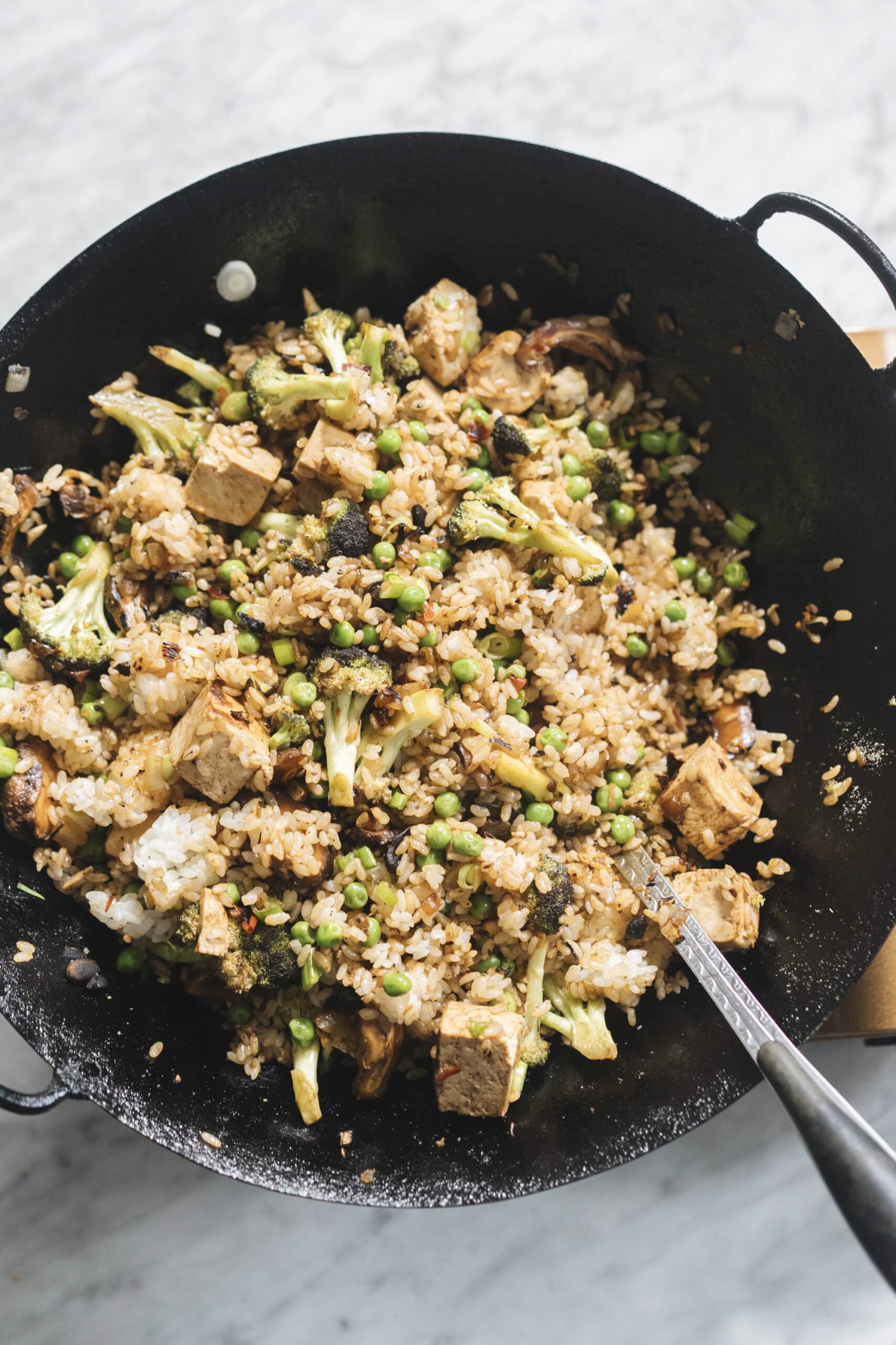 A wok filled with fried rice, tofu cubes, peas, broccoli, and other vegetables, with a serving spoon resting inside.