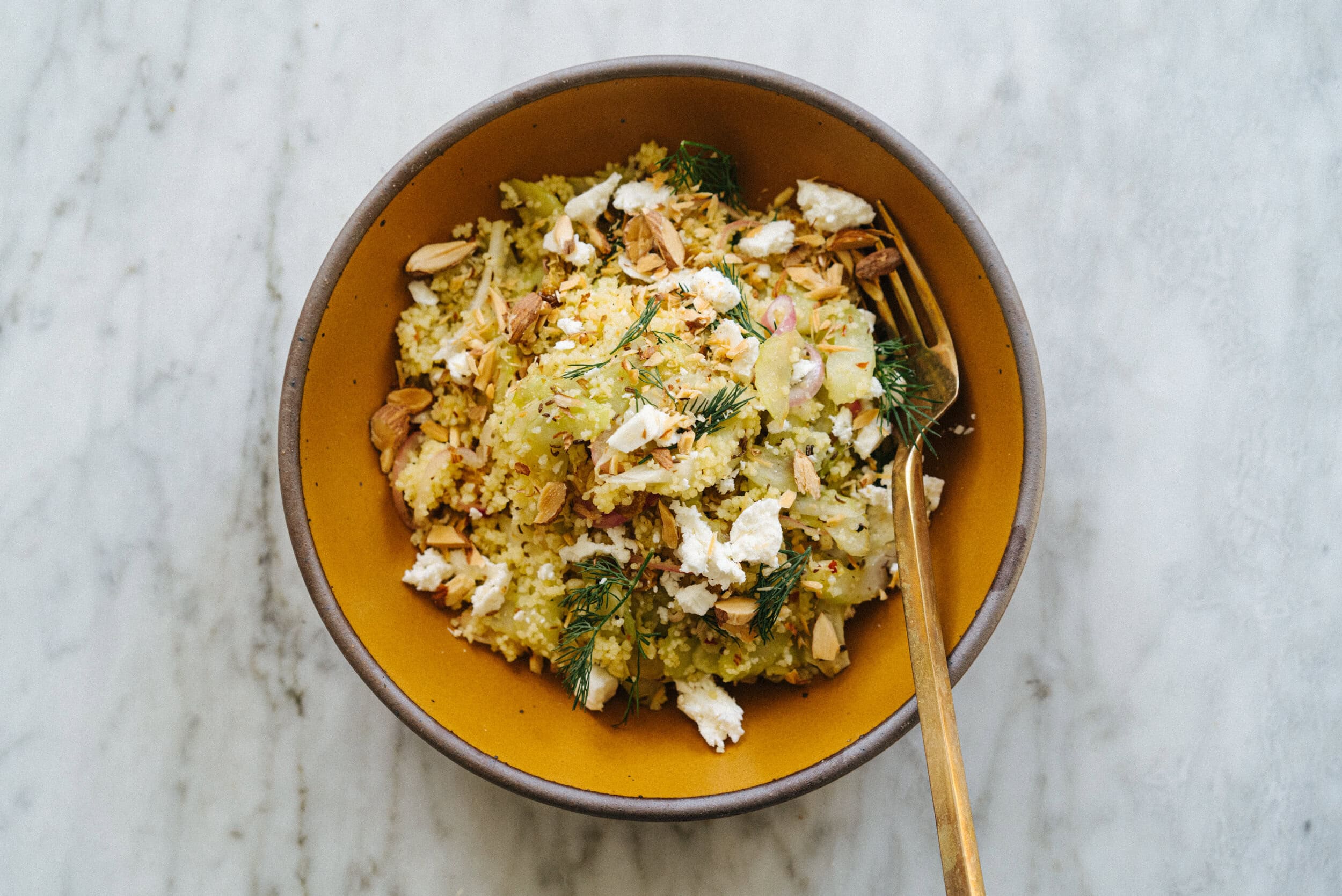 A bowl of couscous salad with herbs, crumbled cheese, chopped nuts, and a gold fork on a marble surface.