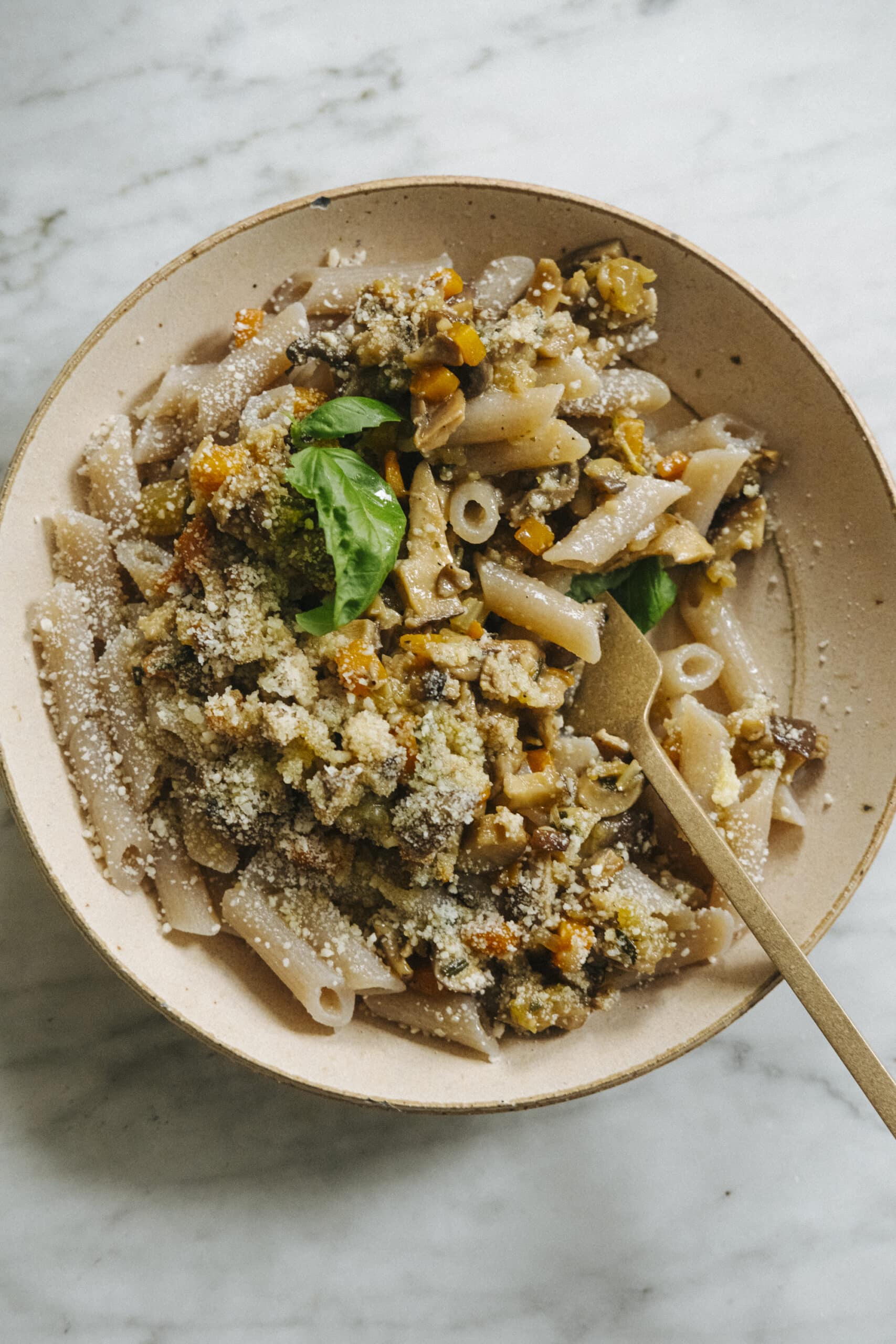 A beige bowl of penne pasta topped with a chunky vegetable sauce, garnished with basil leaves, and sprinkled with grated cheese. A fork rests in the bowl.