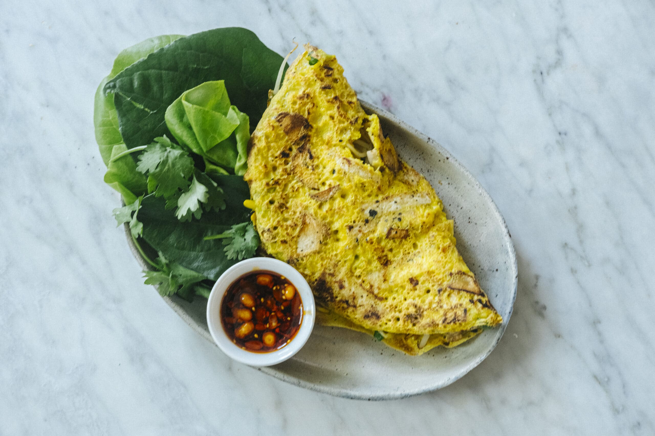 Oval plate with a folded yellow omelette, fresh green leafy herbs, and a small bowl of dipping sauce, on a marble surface.