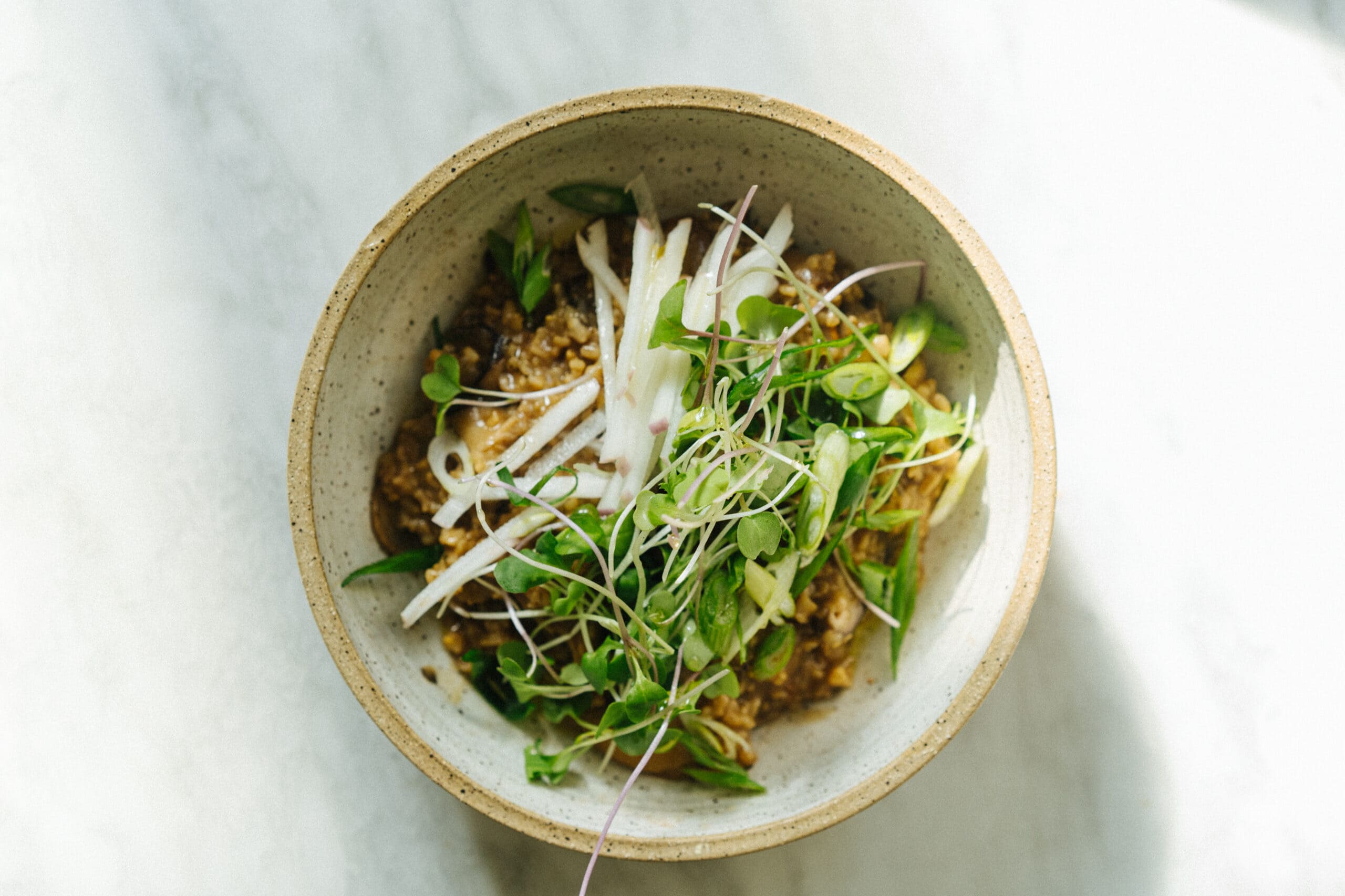 A ceramic bowl filled with a grain-based dish, topped with microgreens and thinly sliced white vegetables, sits on a light-colored surface.