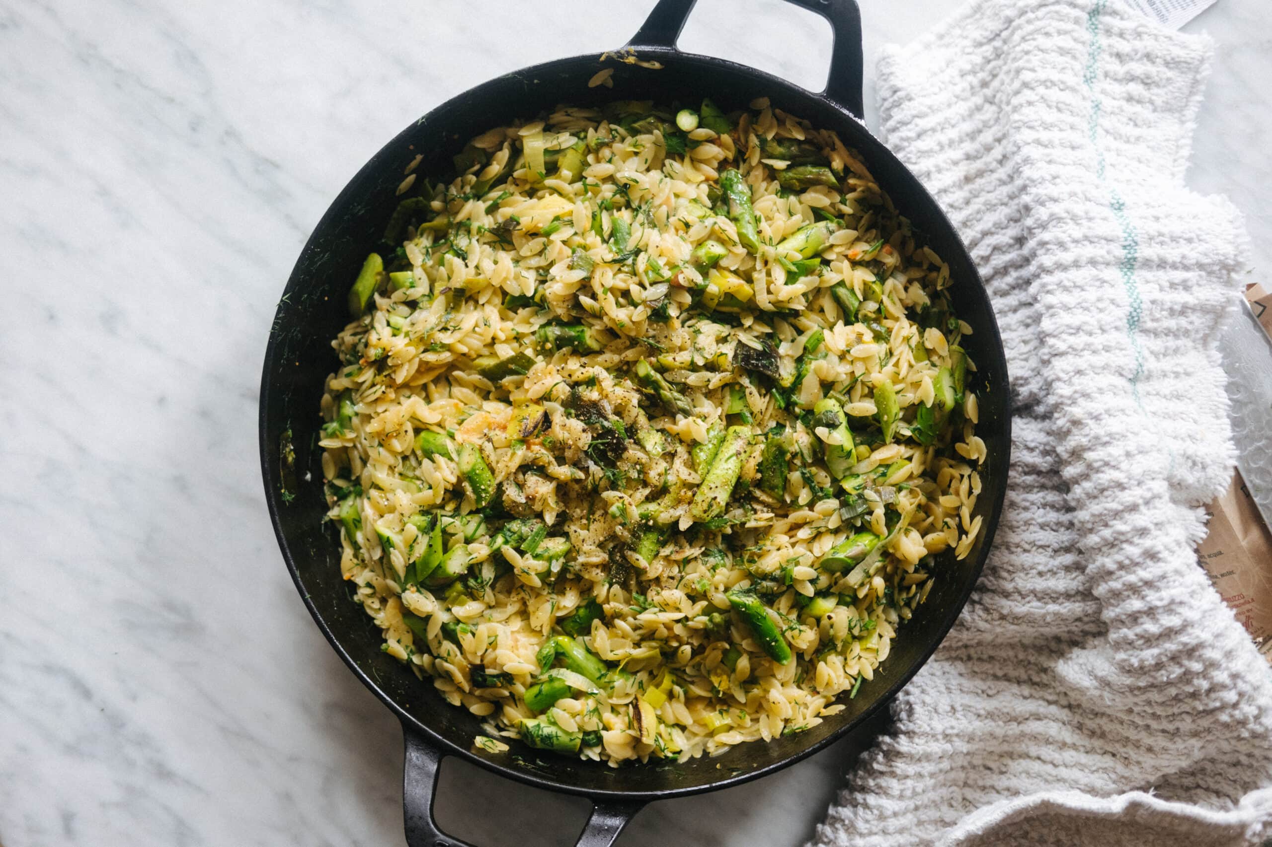 A black skillet filled with cooked orzo pasta mixed with green vegetables on a marble surface next to a textured white towel.