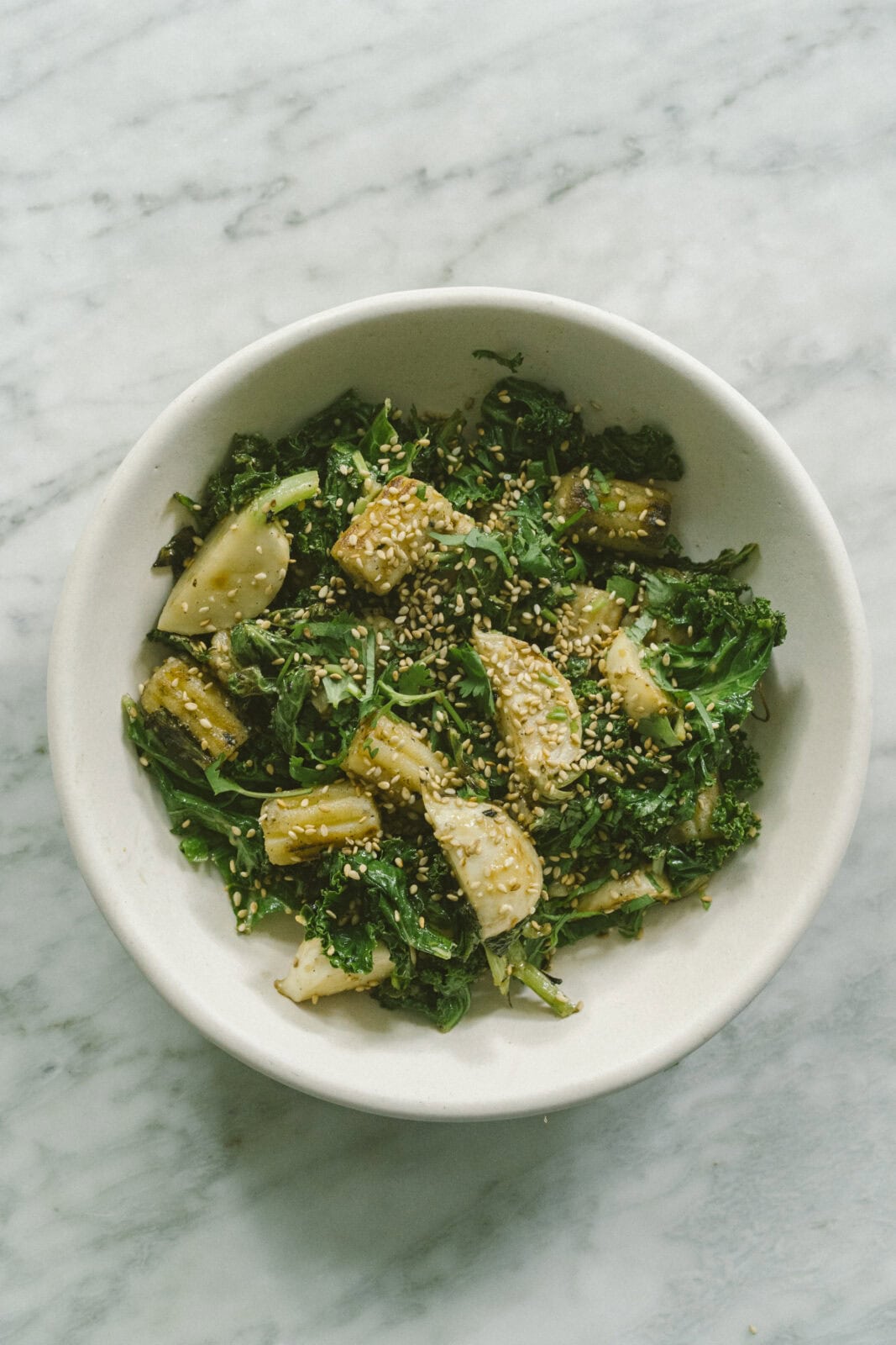 A white bowl filled with cooked leafy greens, chunks of tofu, and sprinkled with sesame seeds on a marble surface.