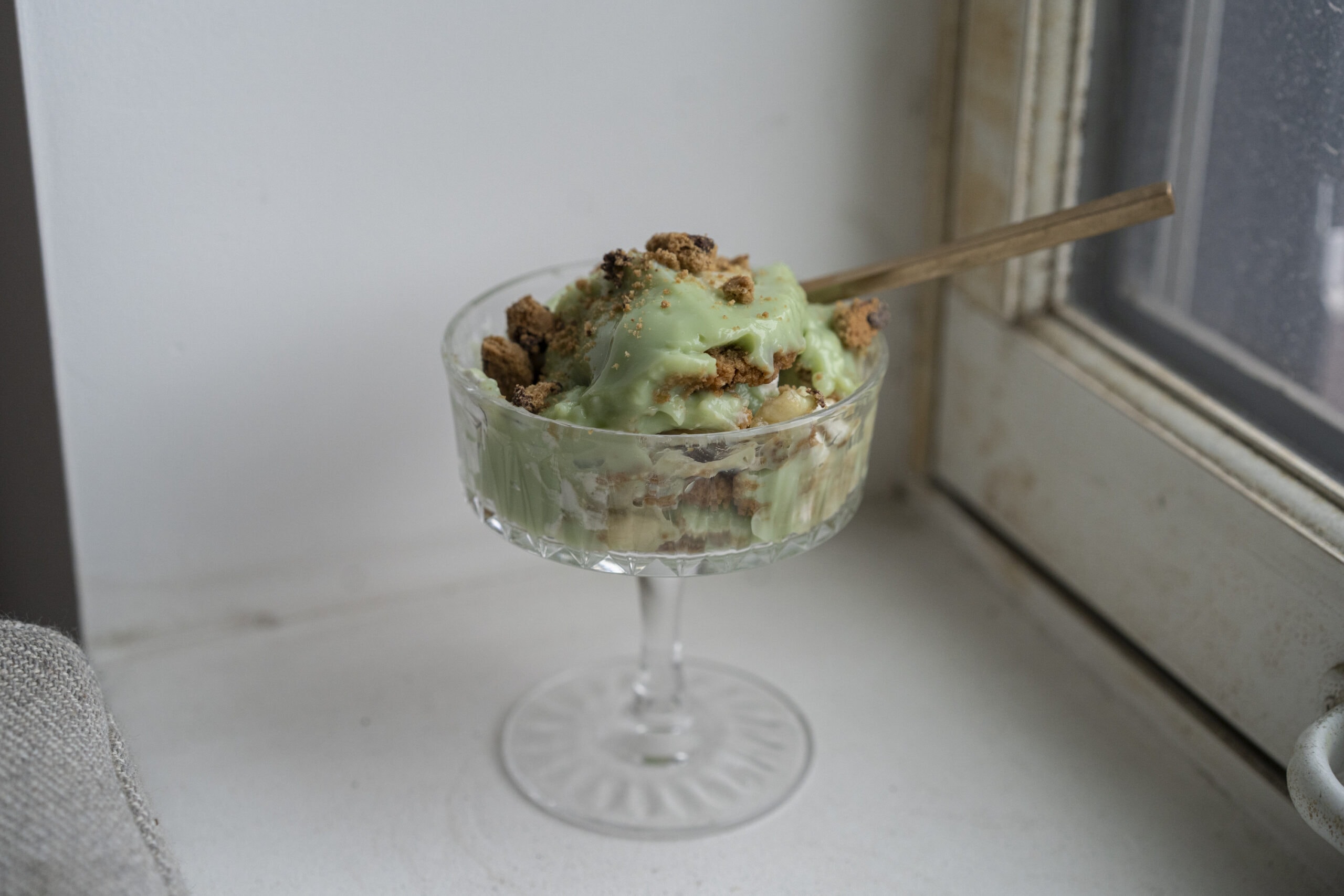 A glass dessert dish filled with light green pudding topped with crumbled cookies, placed on a windowsill with a wooden spoon.