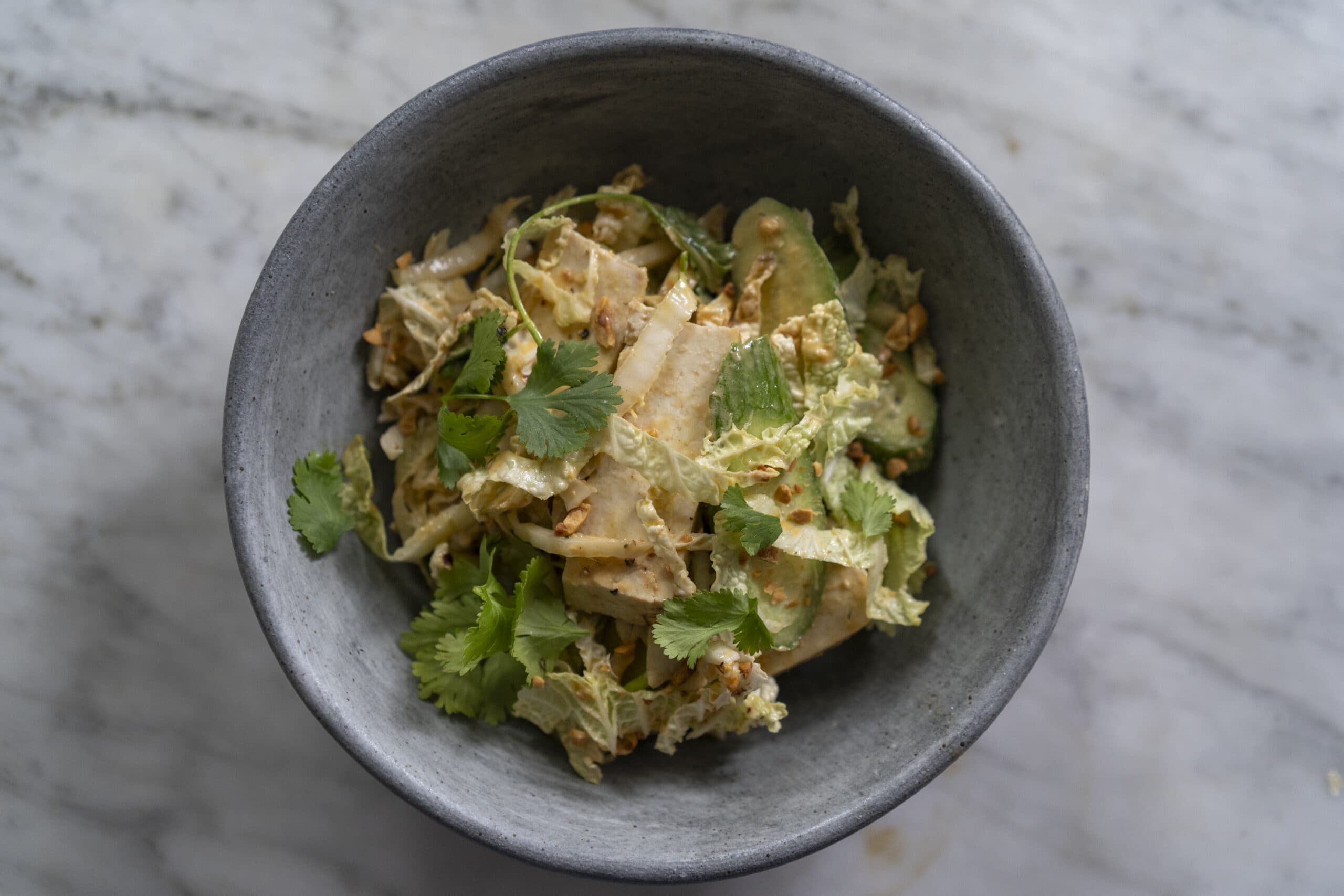 A grey bowl filled with a mixed salad containing sliced vegetables, fresh cilantro leaves, nuts, and a creamy dressing, placed on a marble surface.