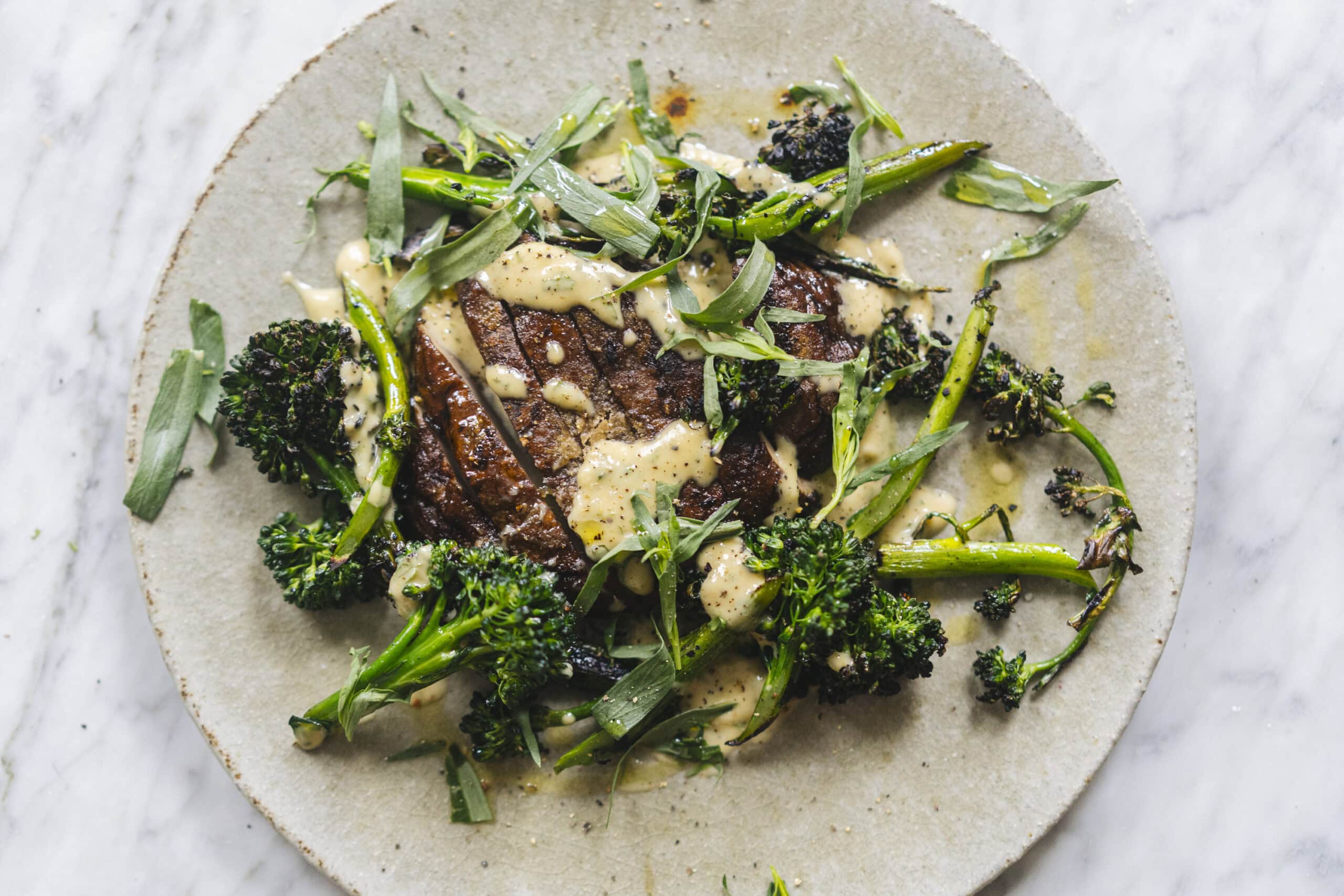 A plate with grilled steak, broccolini, and fresh herbs, topped with a creamy sauce, on a light-colored surface.