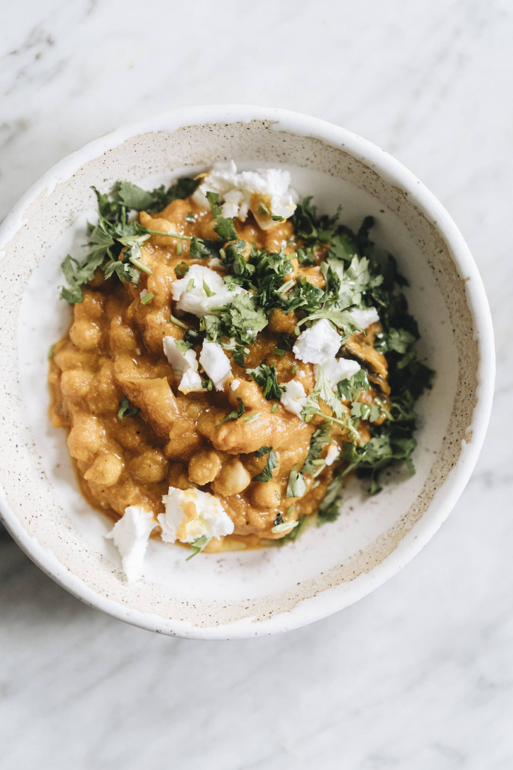 A bowl of chickpea curry topped with crumbled cheese and chopped fresh cilantro, served in a white ceramic bowl on a light surface.