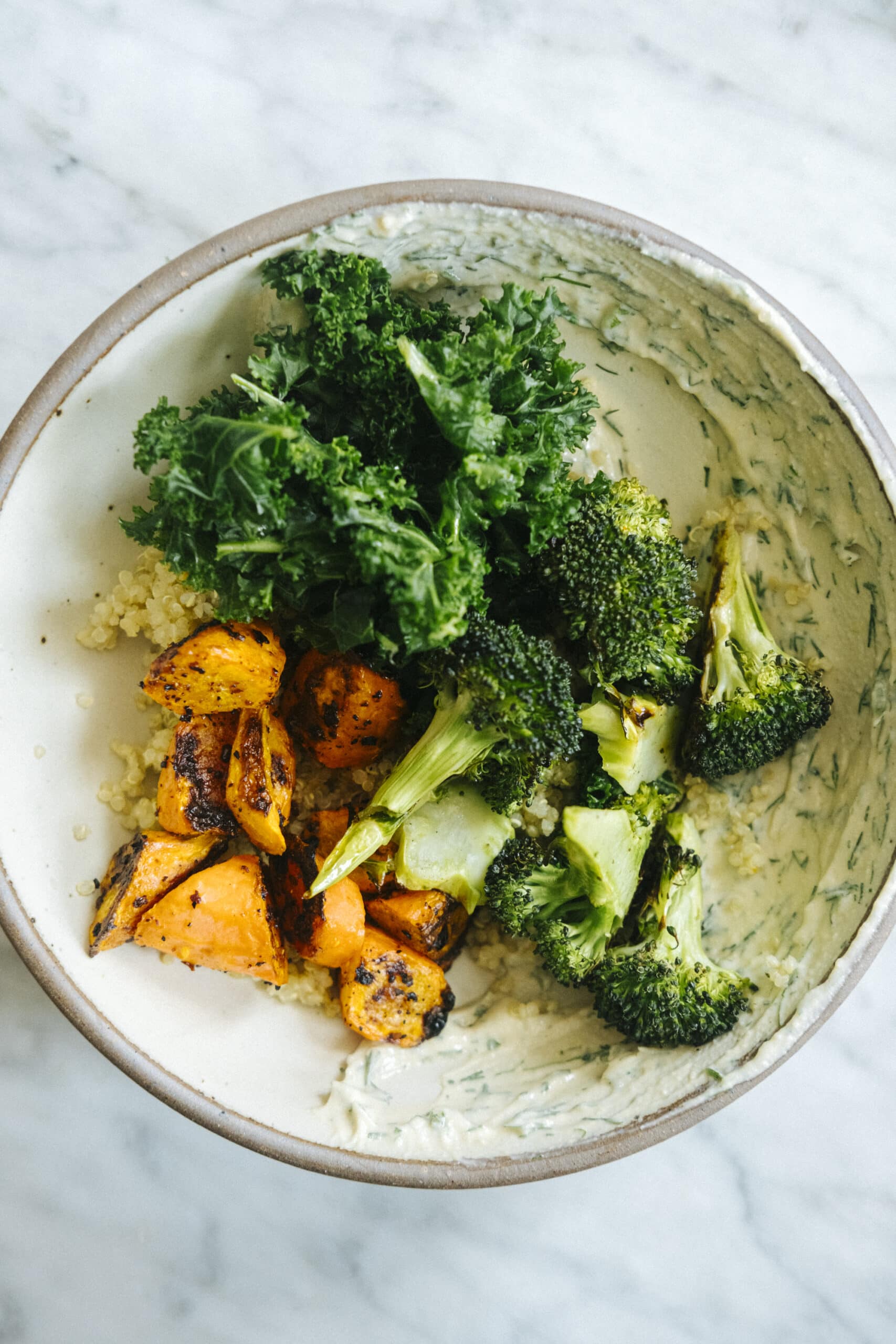 A bowl containing quinoa, roasted sweet potatoes, broccoli, and fresh kale on a marble surface.